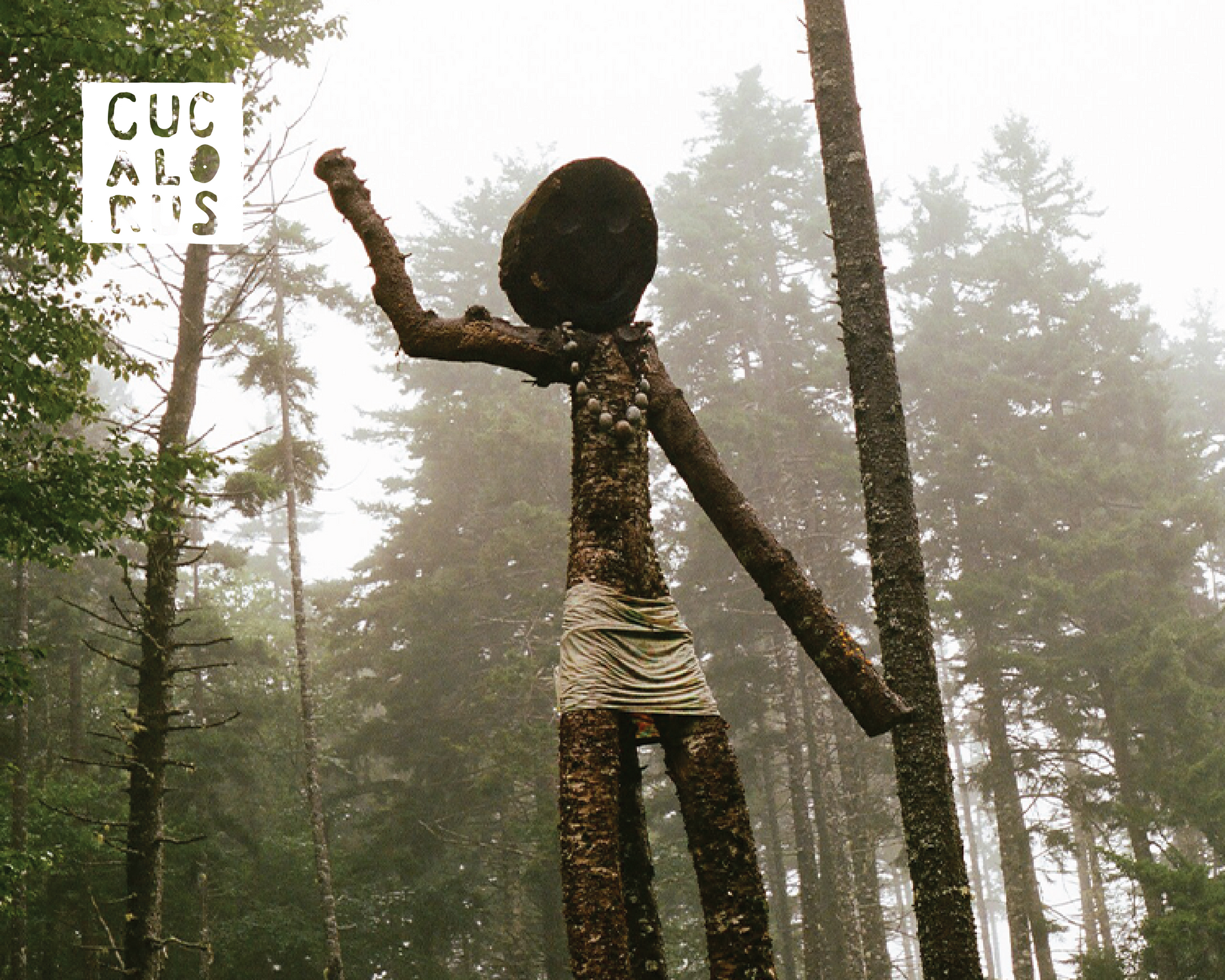 Tall wooden humanoid sculpture in a foggy forest, arm raised beside a trail sign.