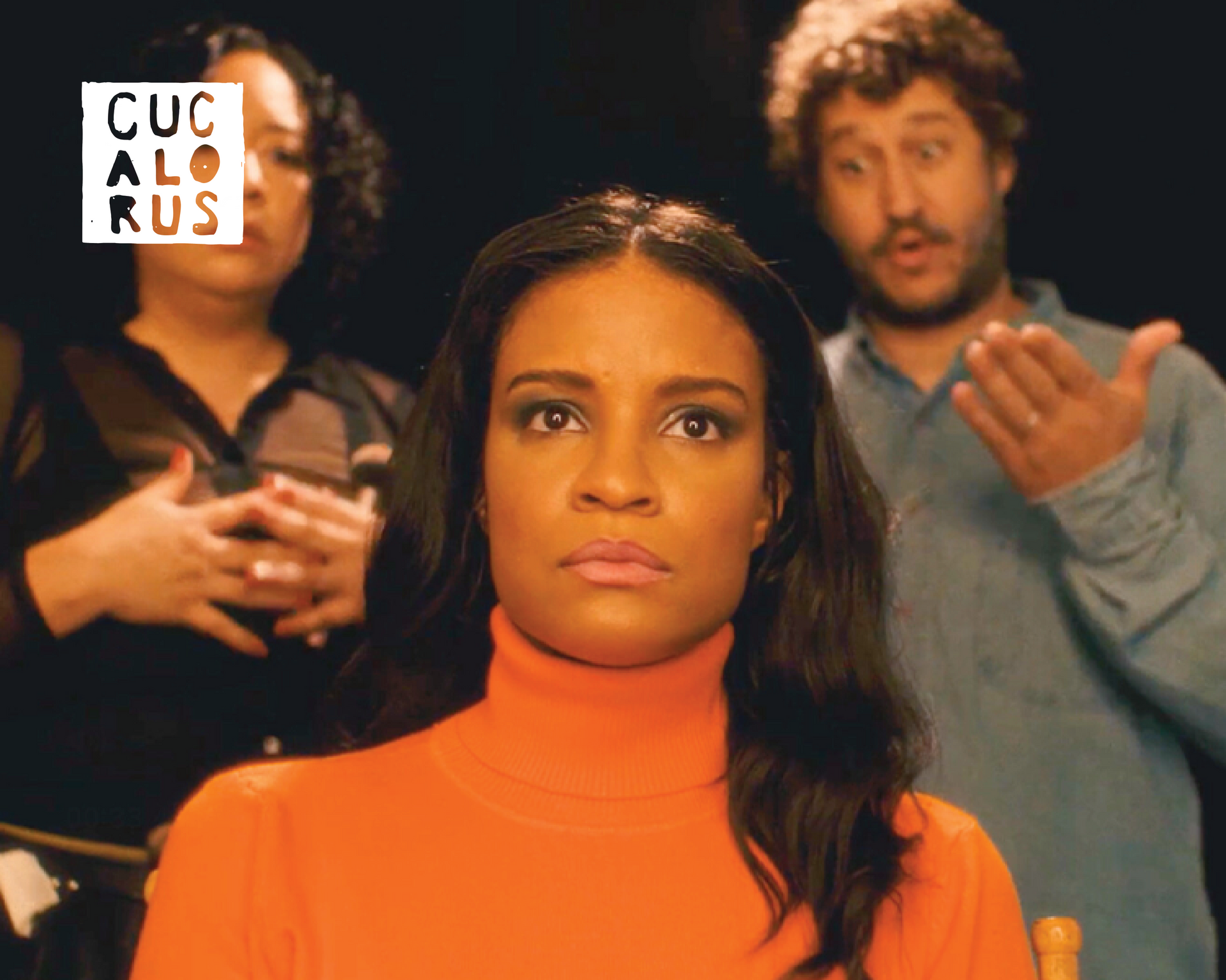 Woman in orange turtleneck centered, with two blurred people behind her and a small logo in the top-left.