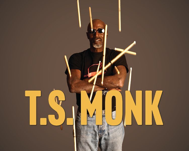 T.S. Monk standing with crossed arms amid glowing drumsticks on a dark background