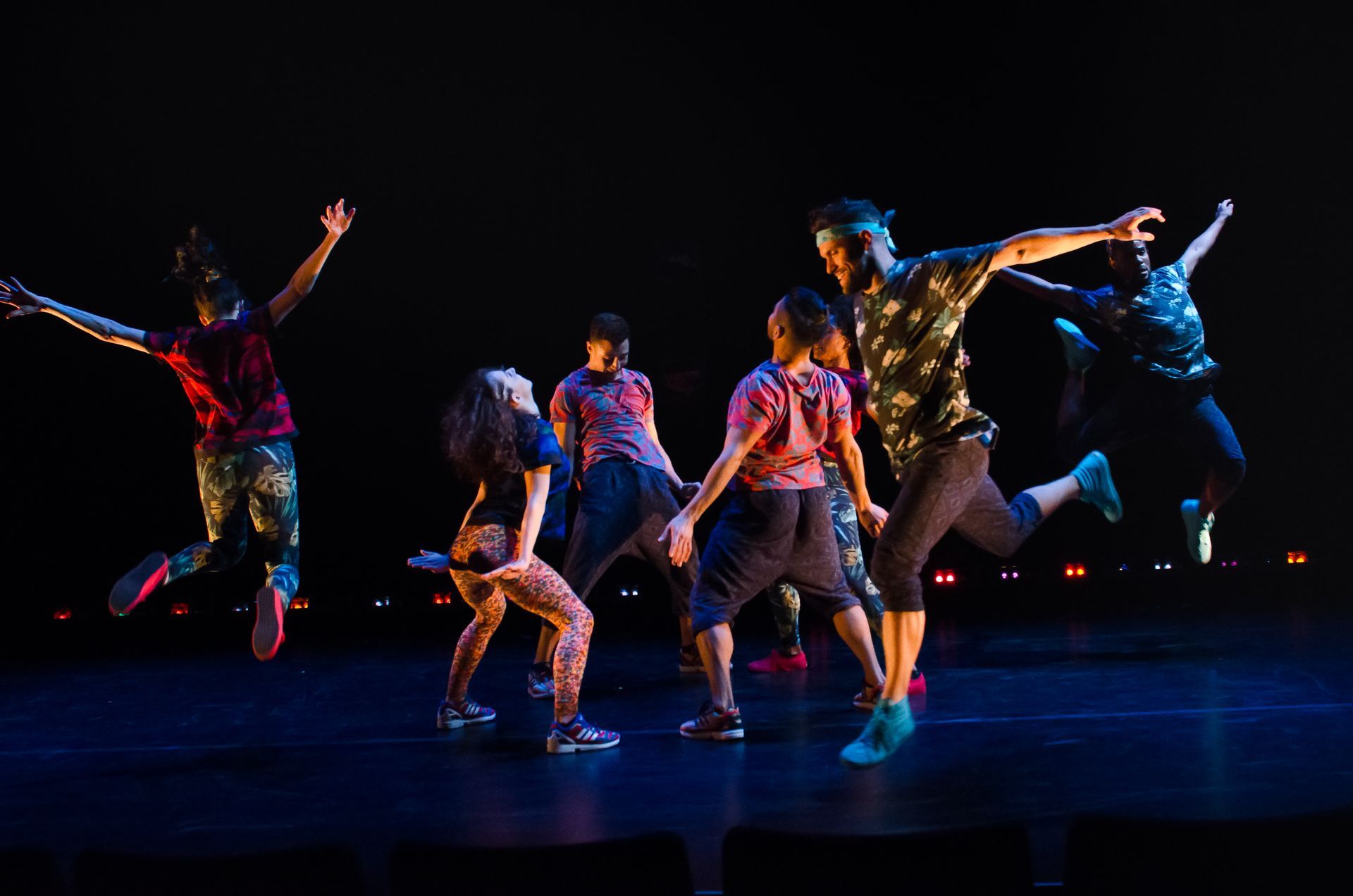 Five dancers mid-jump on a dark stage, lit by colorful spotlights, performing a dynamic routine