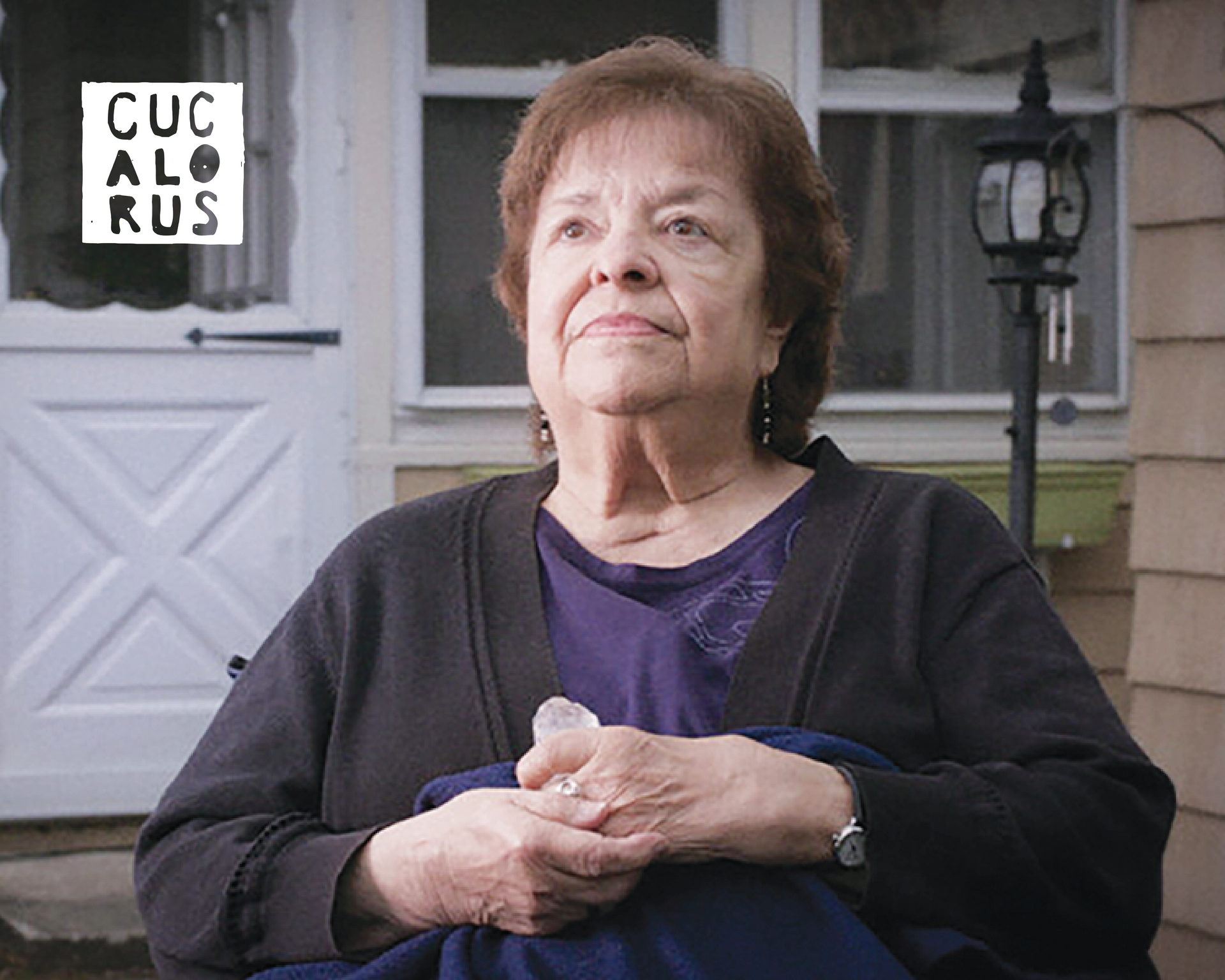 Woman seated outdoors in front of a house, wearing a dark cardigan and looking slightly upward.