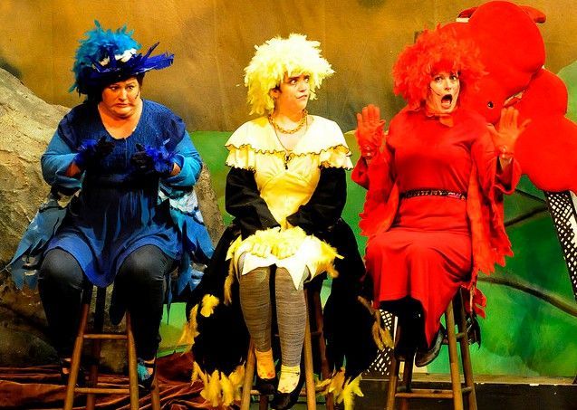 Four performers in colorful costumes sitting on chairs against a painted stage backdrop