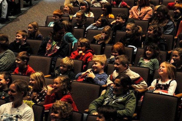 Audience of children seated in theater chairs, watching an event in a dimly lit auditorium