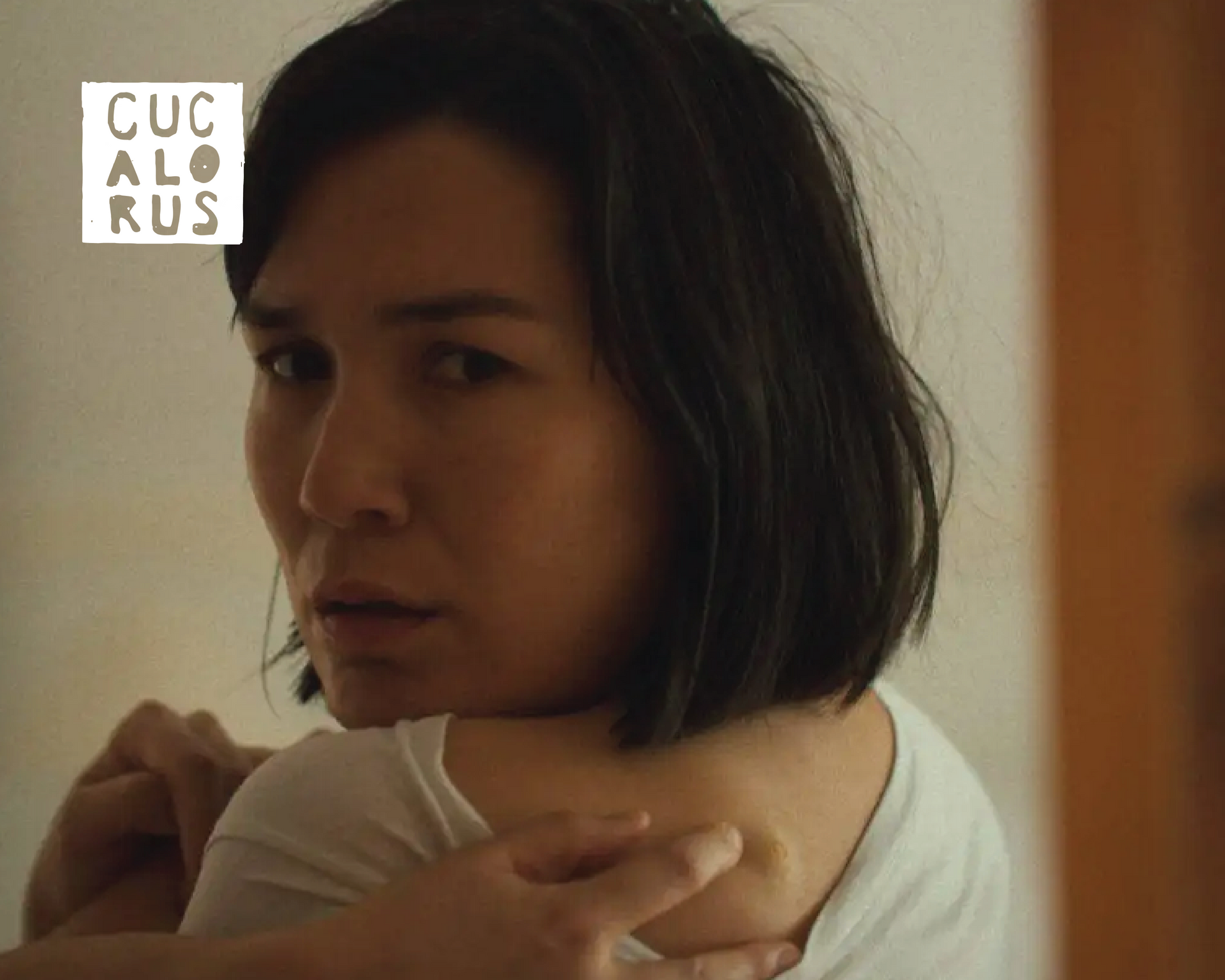 Woman with short dark hair hugging herself indoors, looking toward the camera with a pensive expression.