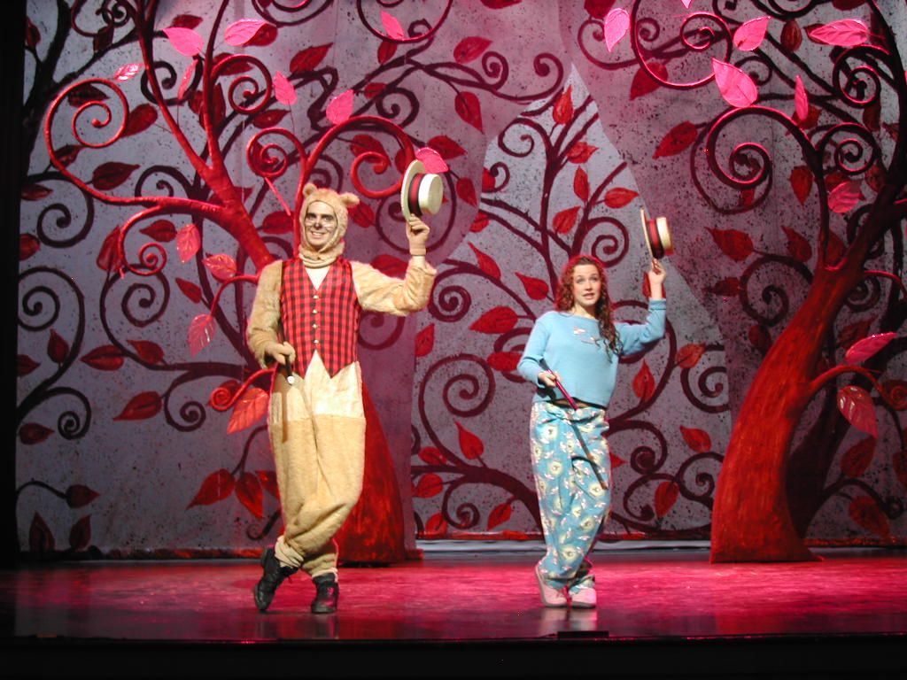 Two performers onstage in colorful costumes, posing under red swirled tree backdrops.
