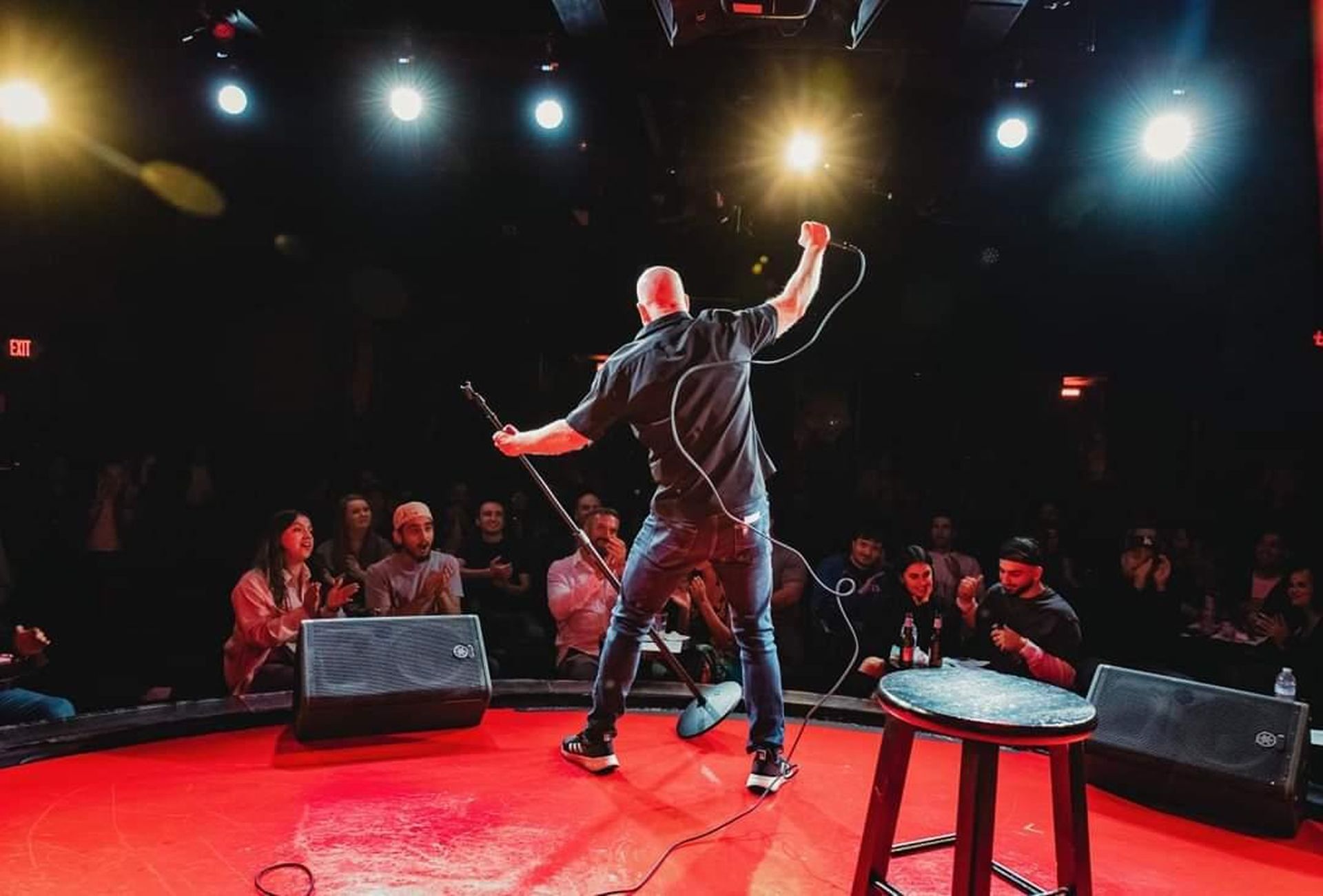 Comedian on a red-lit stage performing to an audience under bright spotlights