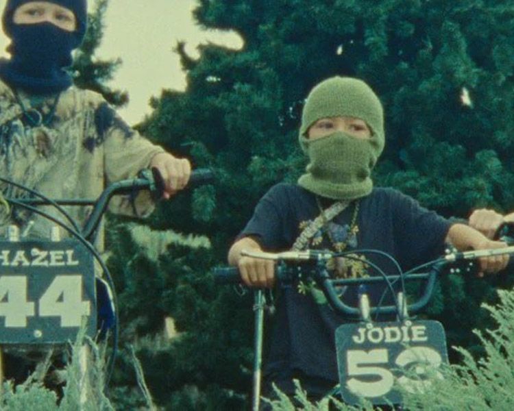 Two masked children on bicycles, one labeled Hazel 44 and the other Jodie 53, outdoors among trees.