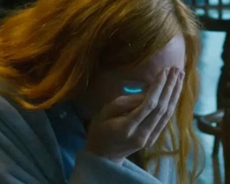 A red-haired person covers their face with one hand, glowing blue eye visible in a dim room.