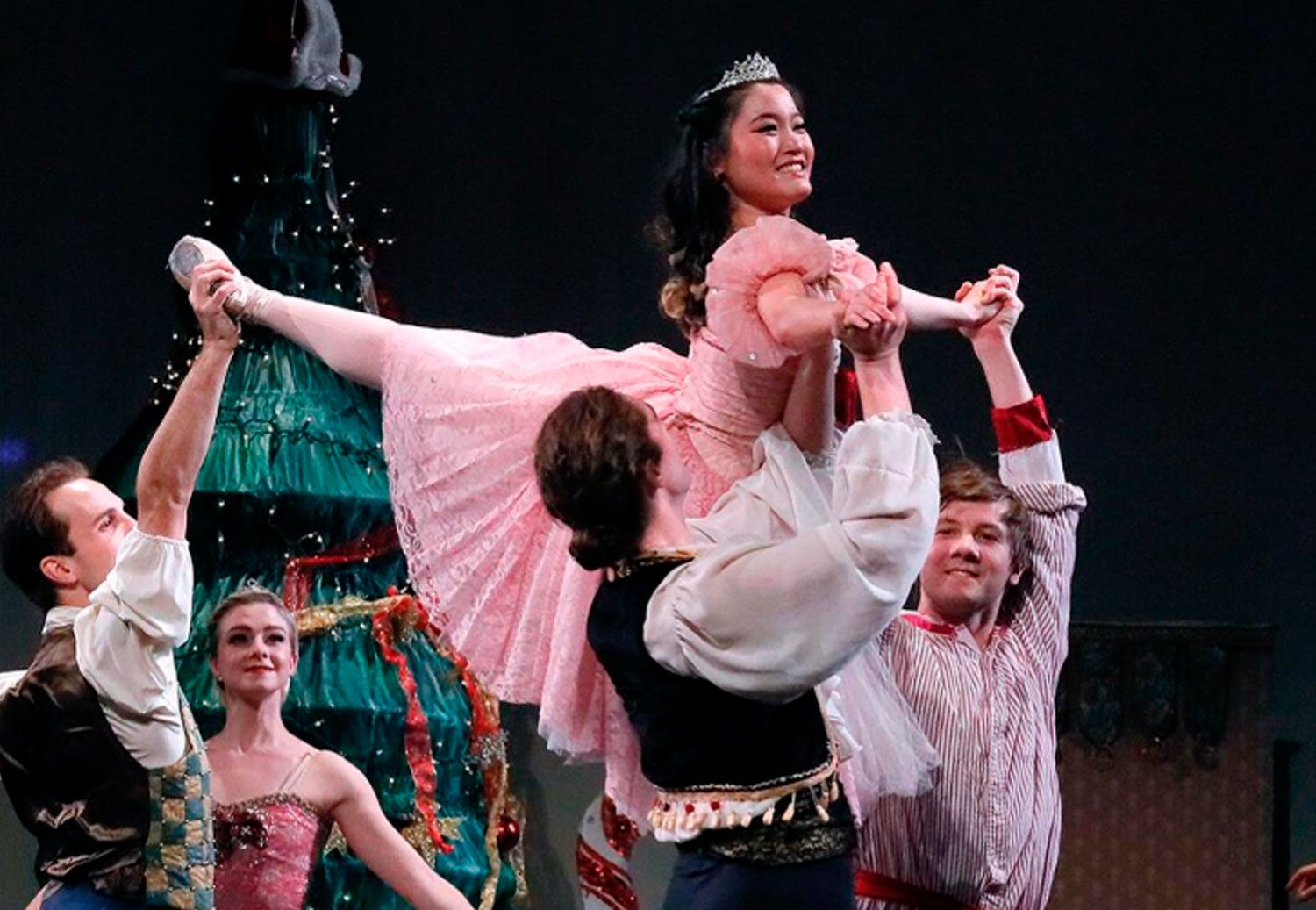 Ballet dancers lift a smiling performer in a pink costume onstage, with a Christmas tree in the background