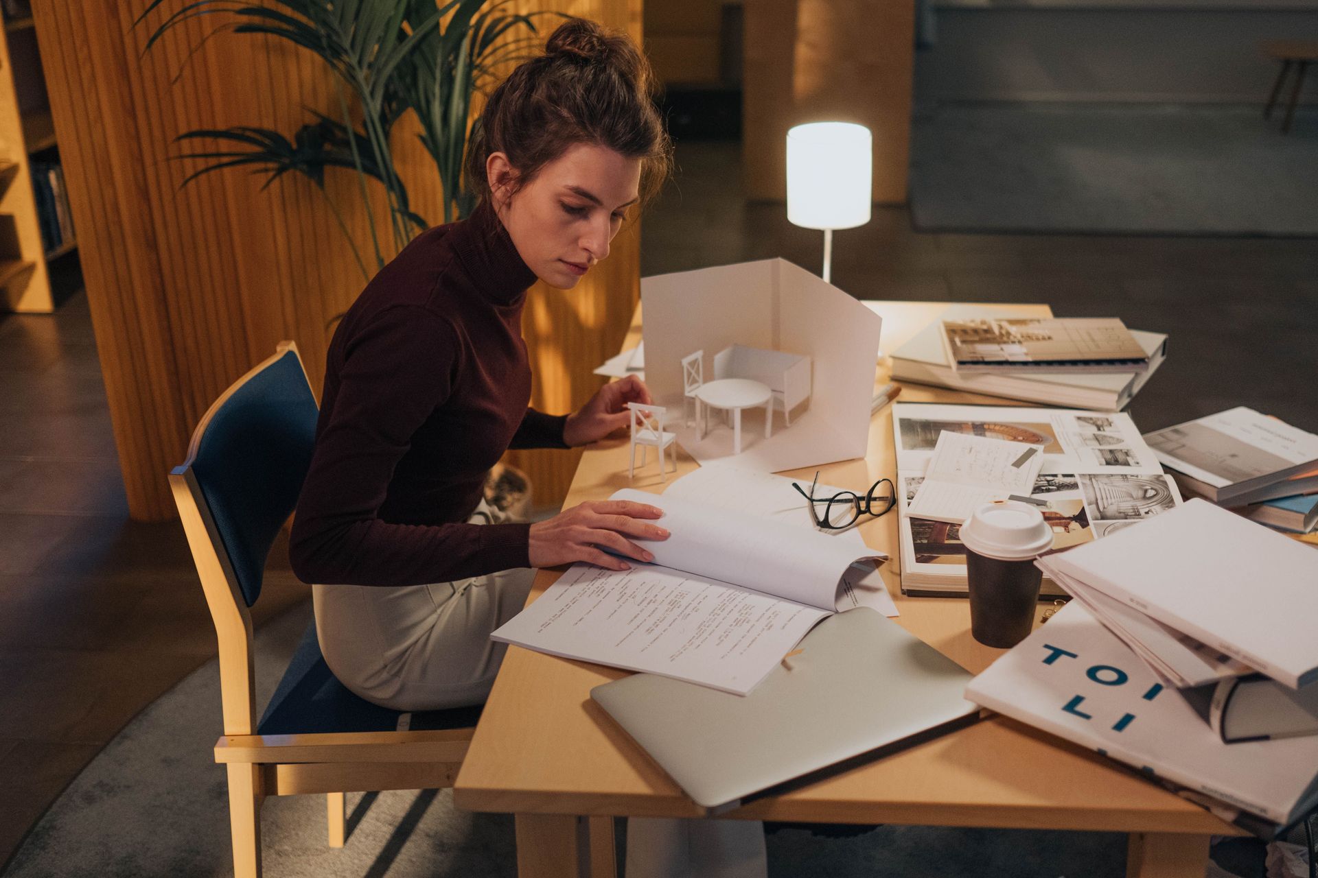 Person working at a cluttered desk with papers and a laptop in a warmly lit office.