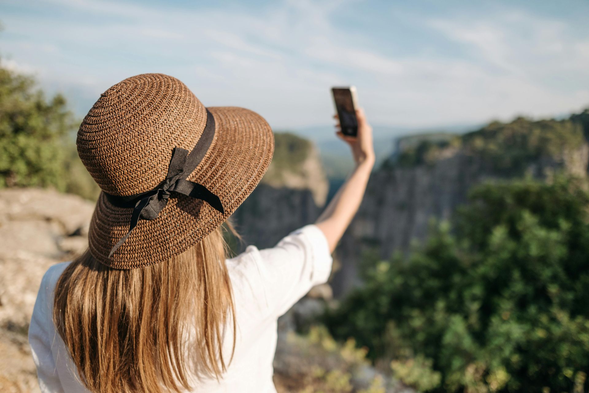 Woman in a straw hat taking a selfie at a scenic cliff overlook under a blue sky