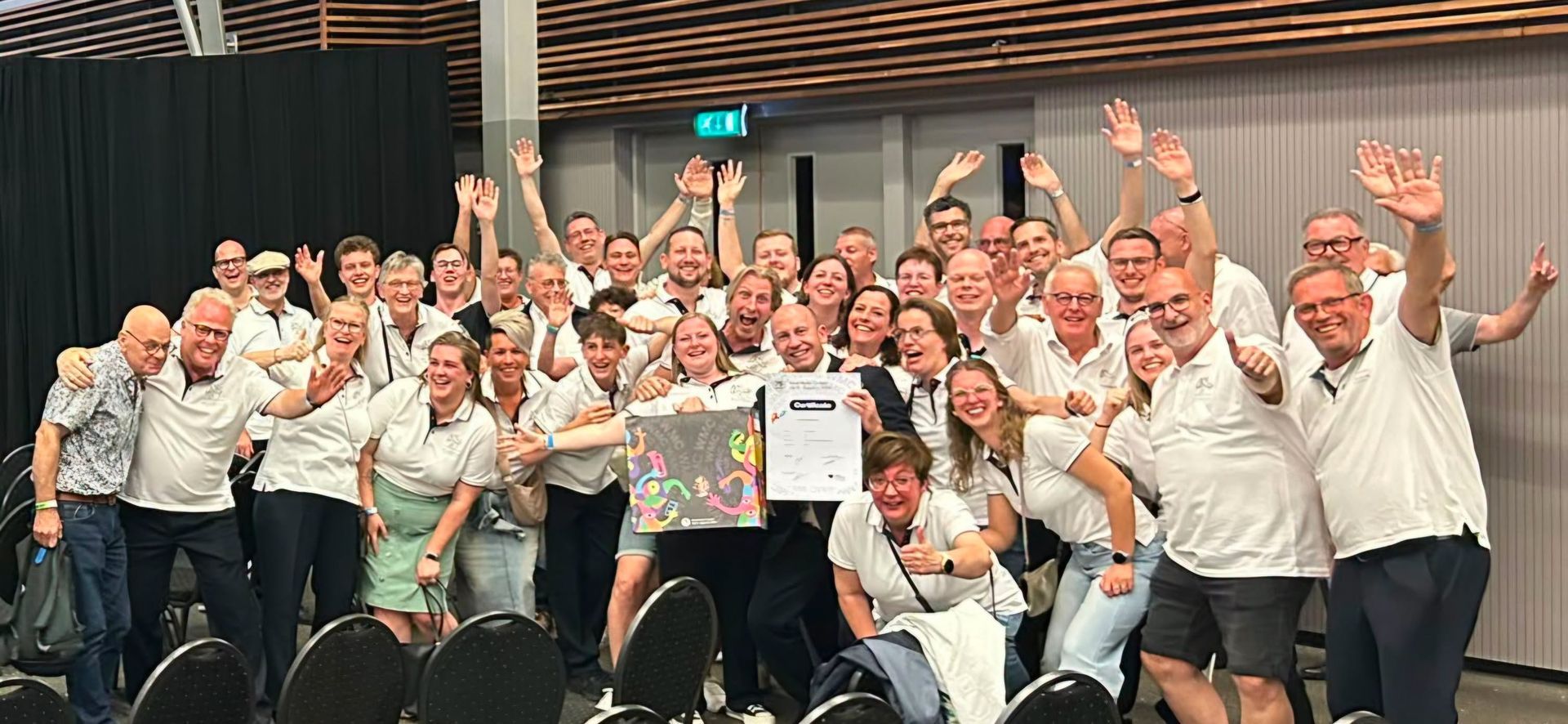 Groep mensen in witte shirts die binnen poseren en zwaaien, feestvierend rond een kleurrijk kaartbord.