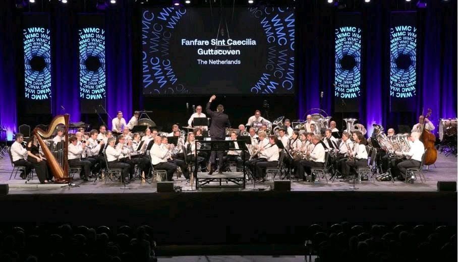 Orkest dat optreedt op een verlicht podium met paarse zuilen en een projectiescherm op de achtergrond