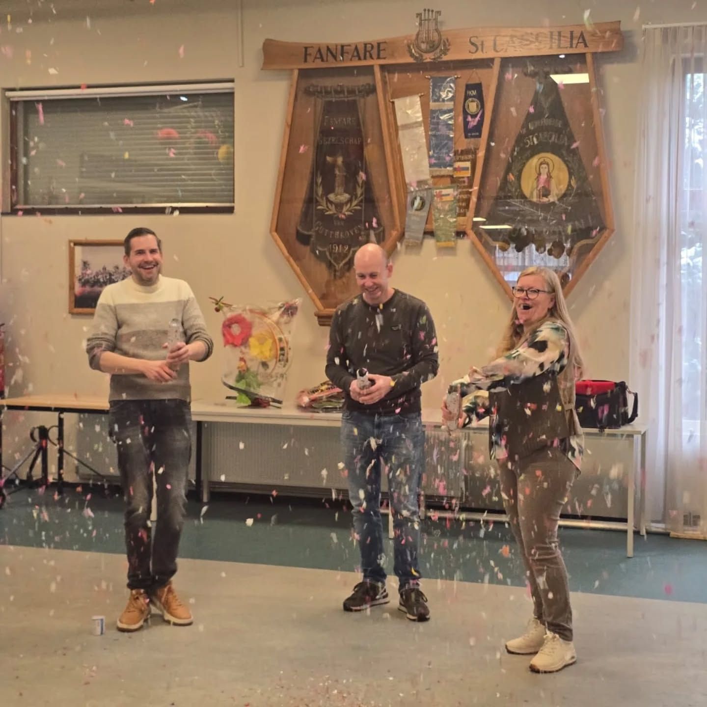 Drie mensen vieren feest met confetti in een kamer; boven hen hangt een spandoek met de tekst