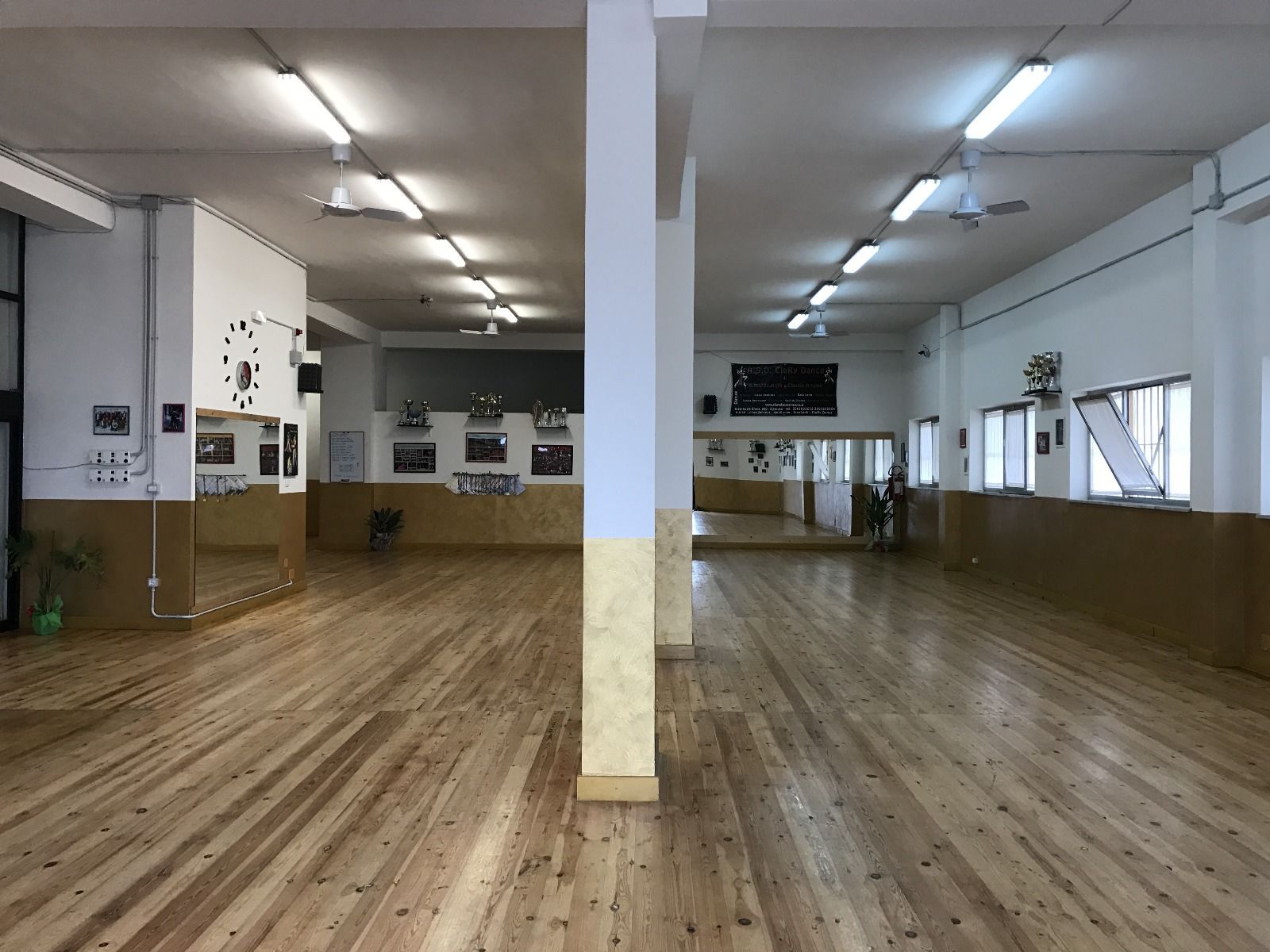 Studio di danza vuoto con pavimento in legno, specchi e una colonna centrale.