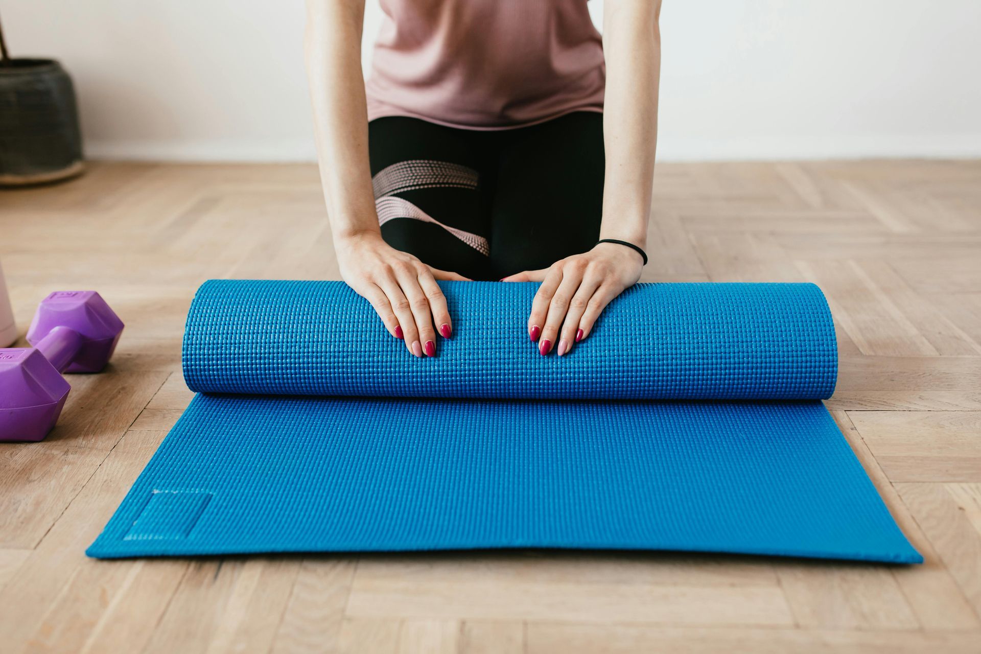 Persona che stende un tappetino da yoga blu su un pavimento in legno, con i manubri visibili.