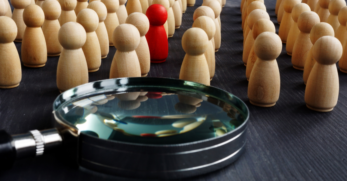 Magnifying glass lying in front of rows of wooden peg people. One peg person is painted red.