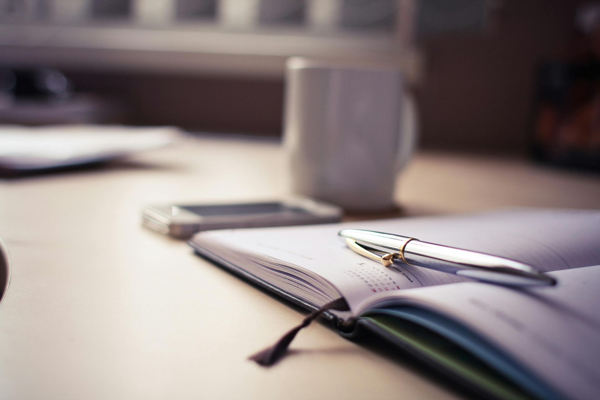 An opened diary planner with a pen on top. There is a mug and mobile phone in the background