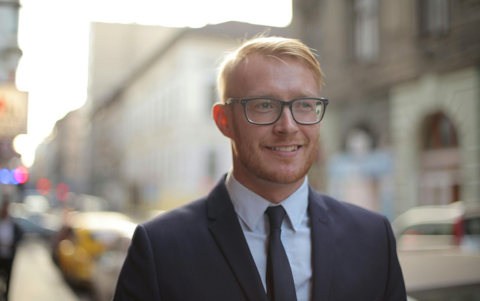 Smiling man in glasses and a navy suit standing on a city street in bright sunlight