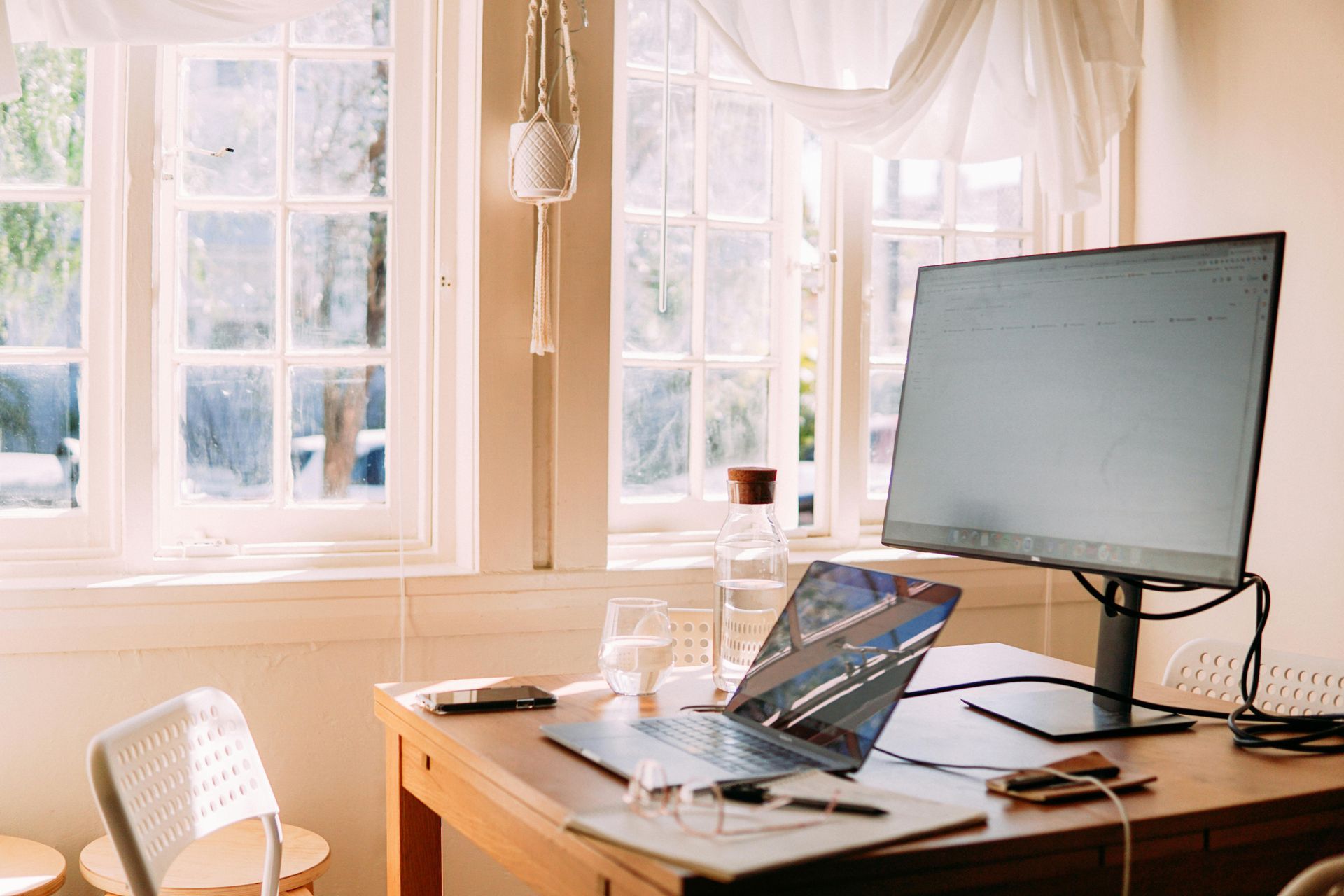 Sunlit home office desk with laptop, large monitor, and window view