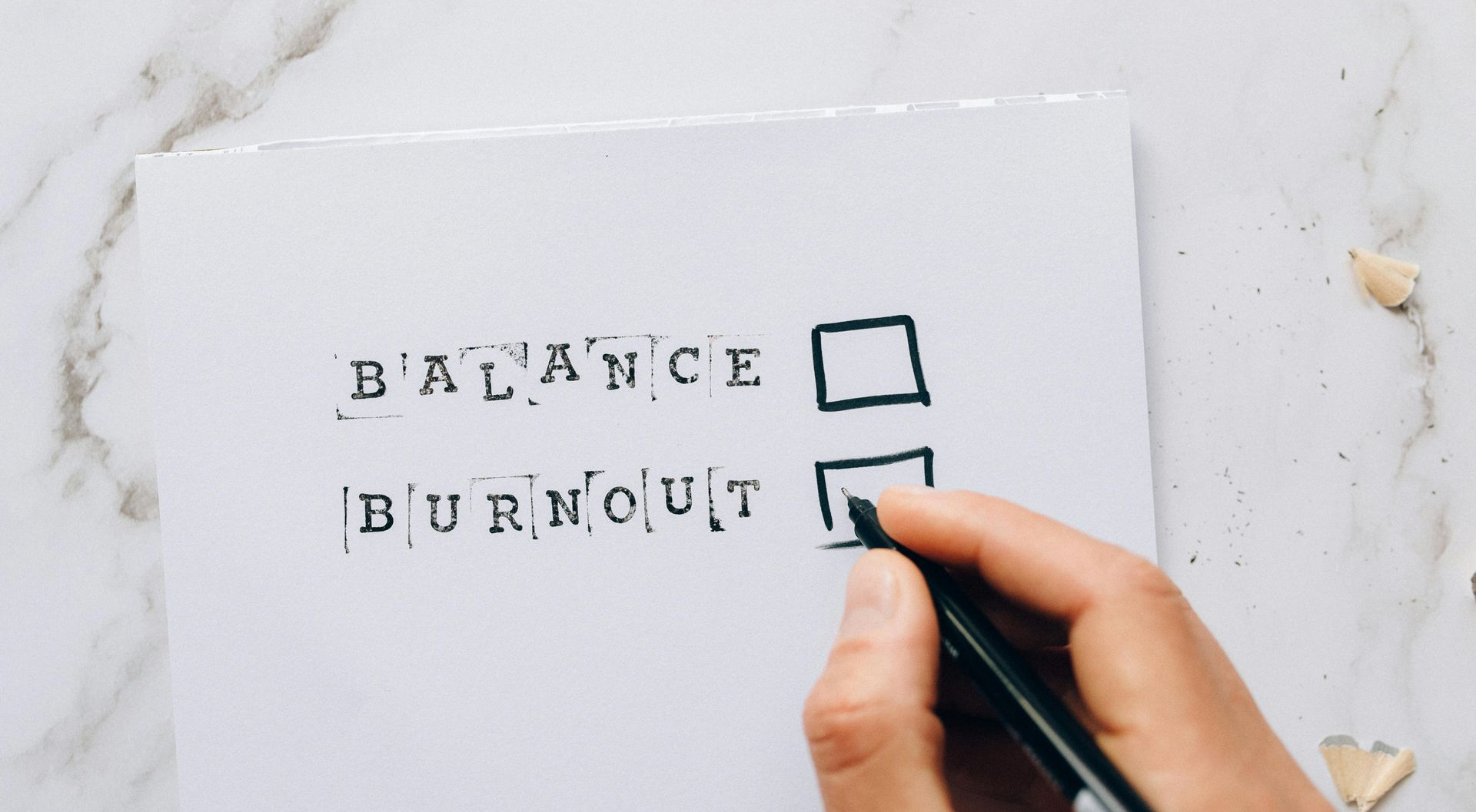 Hand checking the box next to โBURNOUTโ on a checklist beside โBALANCEโ