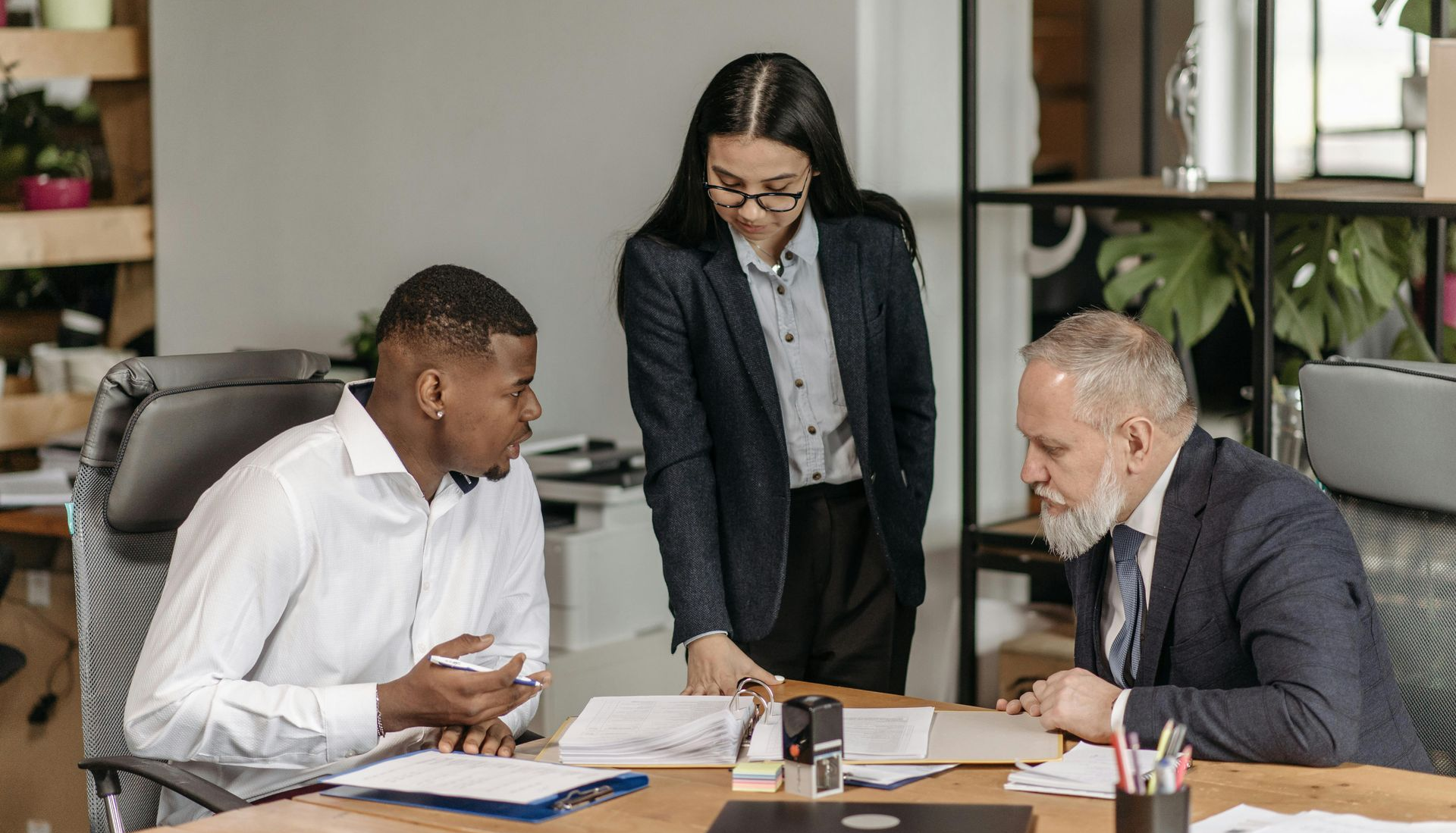 3 office colleagues having a discussion at an office desk. The desk has folders, papers, stationery and a laptop on it