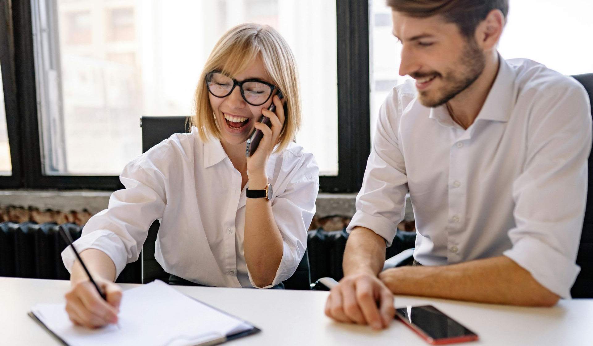 Two coworkers in white shirts smiling at a desk, reviewing papers and talking on a phone in a bright office.