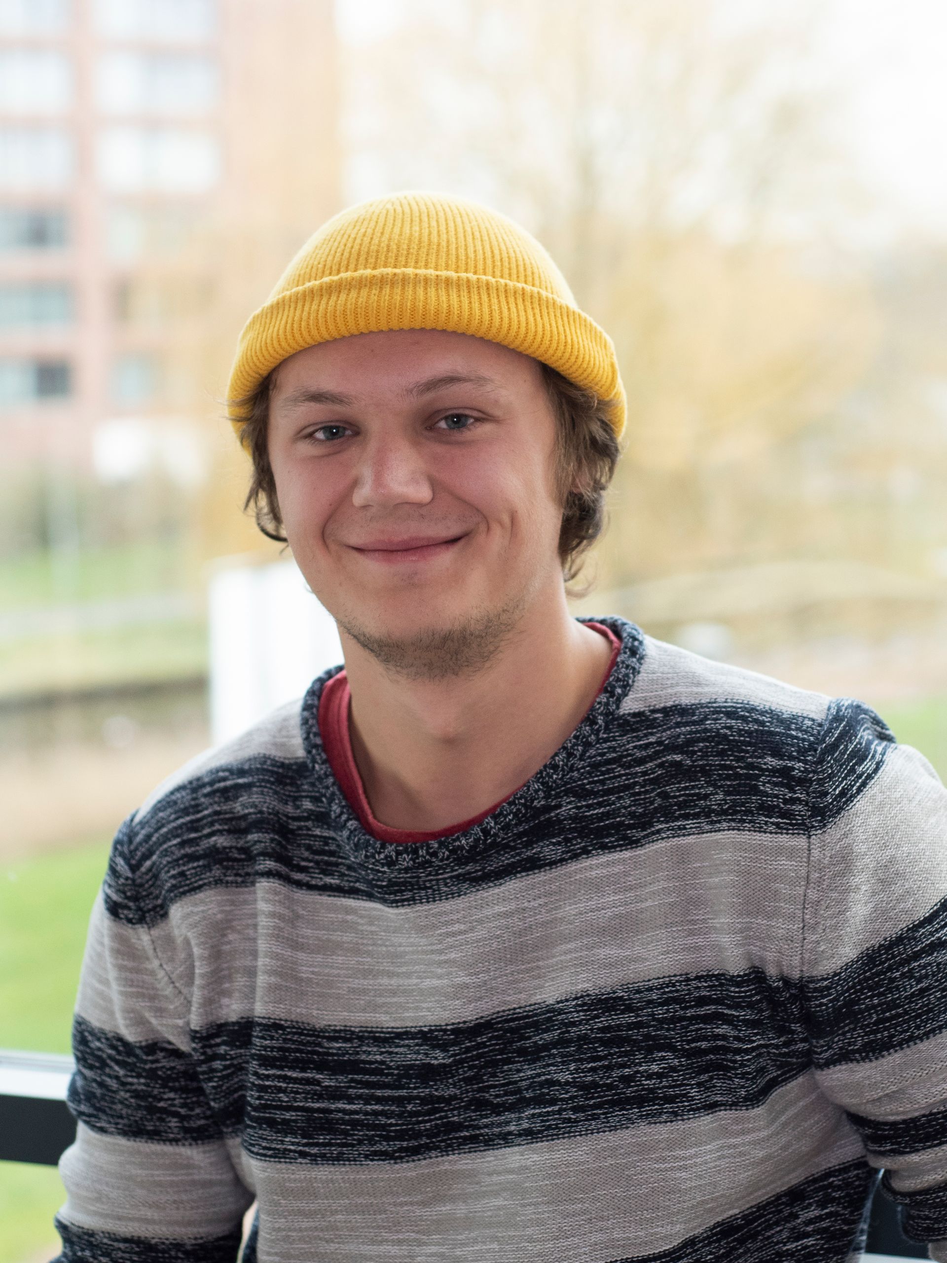 Een jongeman draagt een gele muts en een gestreepte trui en lacht.