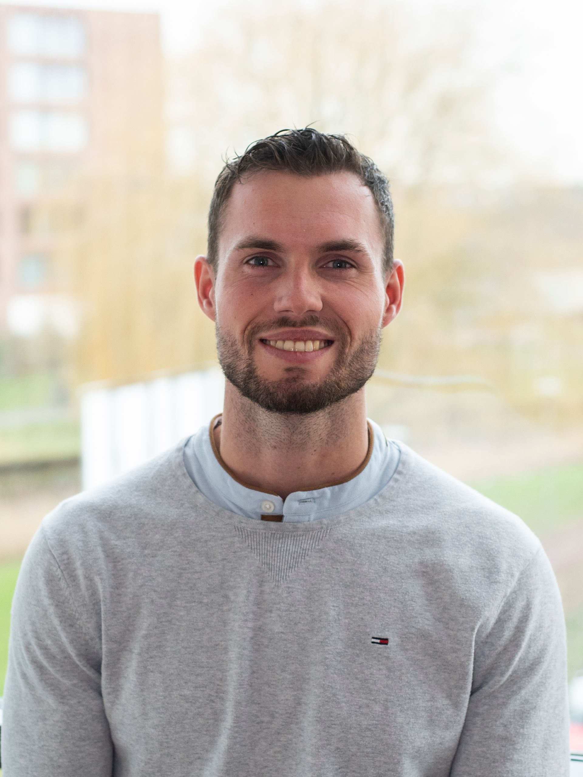Een man met een baard draagt een grijze trui en lacht naar de camera.