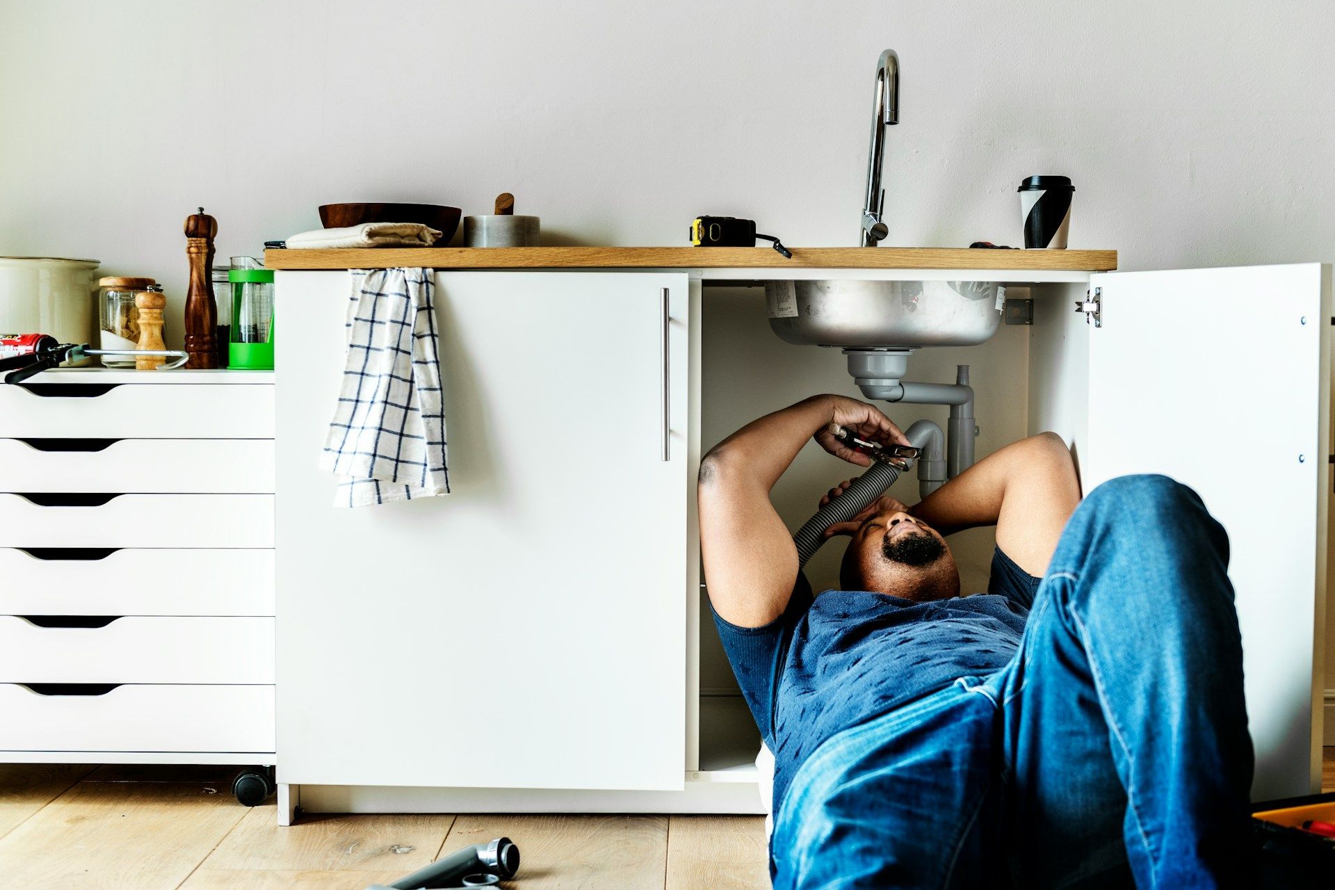 Person lying under a kitchen sink, fixing plumbing with tools on the floor.