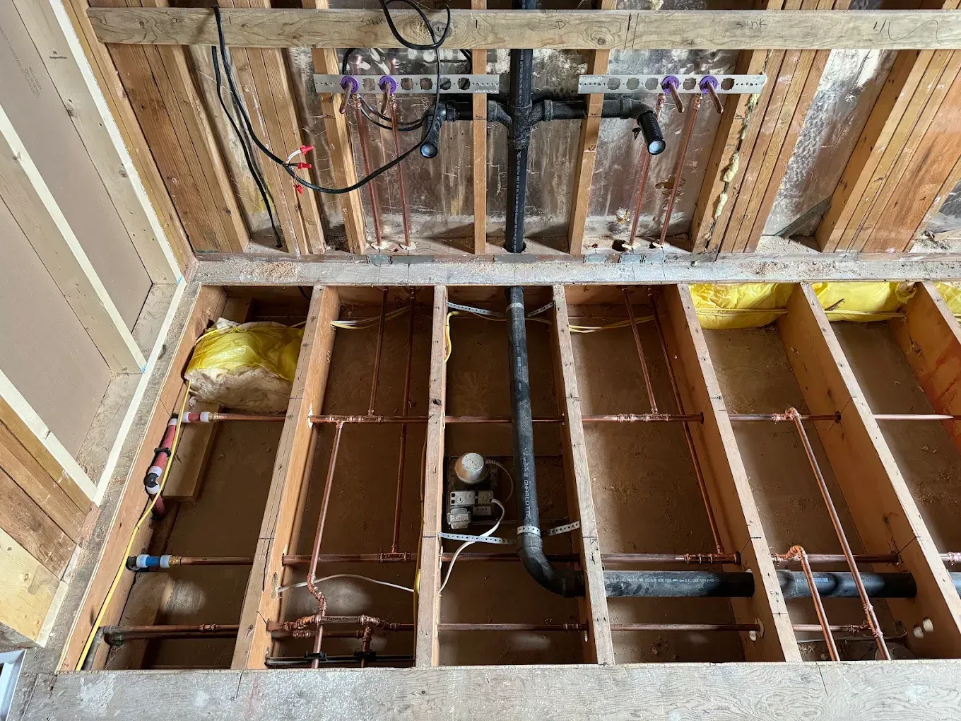 Open wall framing and subfloor joists with exposed plumbing pipes in an unfinished bathroom renovation.