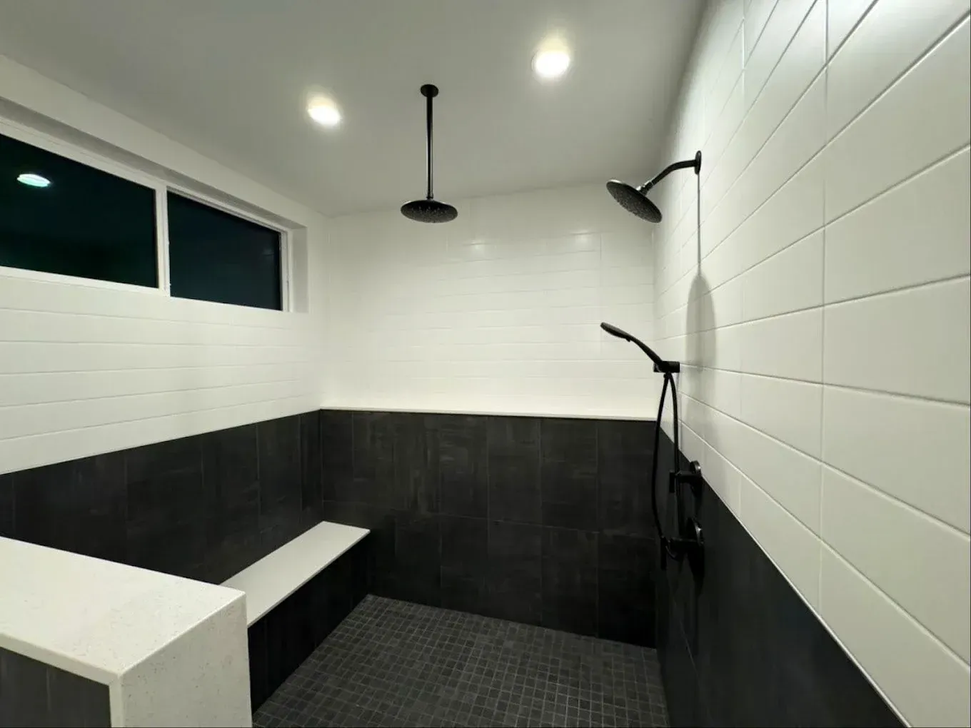 Modern shower room with white and black tile, bench, overhead lights, and multiple showerheads