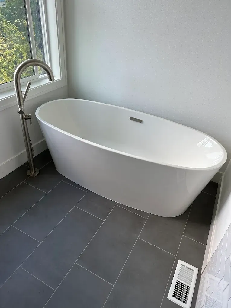 Freestanding white bathtub beside a window in a modern bathroom with dark tile flooring