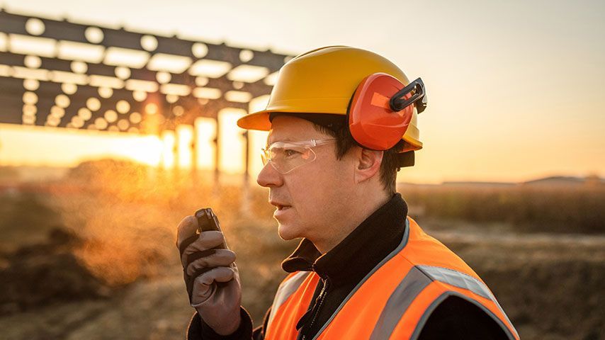 A construction worker wearing a hard hat and ear muffs is talking on a walkie talkie.
