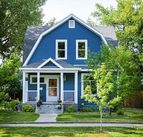 Blue house with white trim, porch, and landscaping; trees surround.