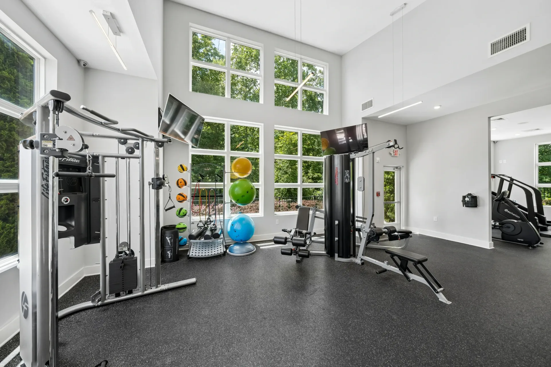 Fitness center with weight machines, cardio equipment, and large windows overlooking trees.
