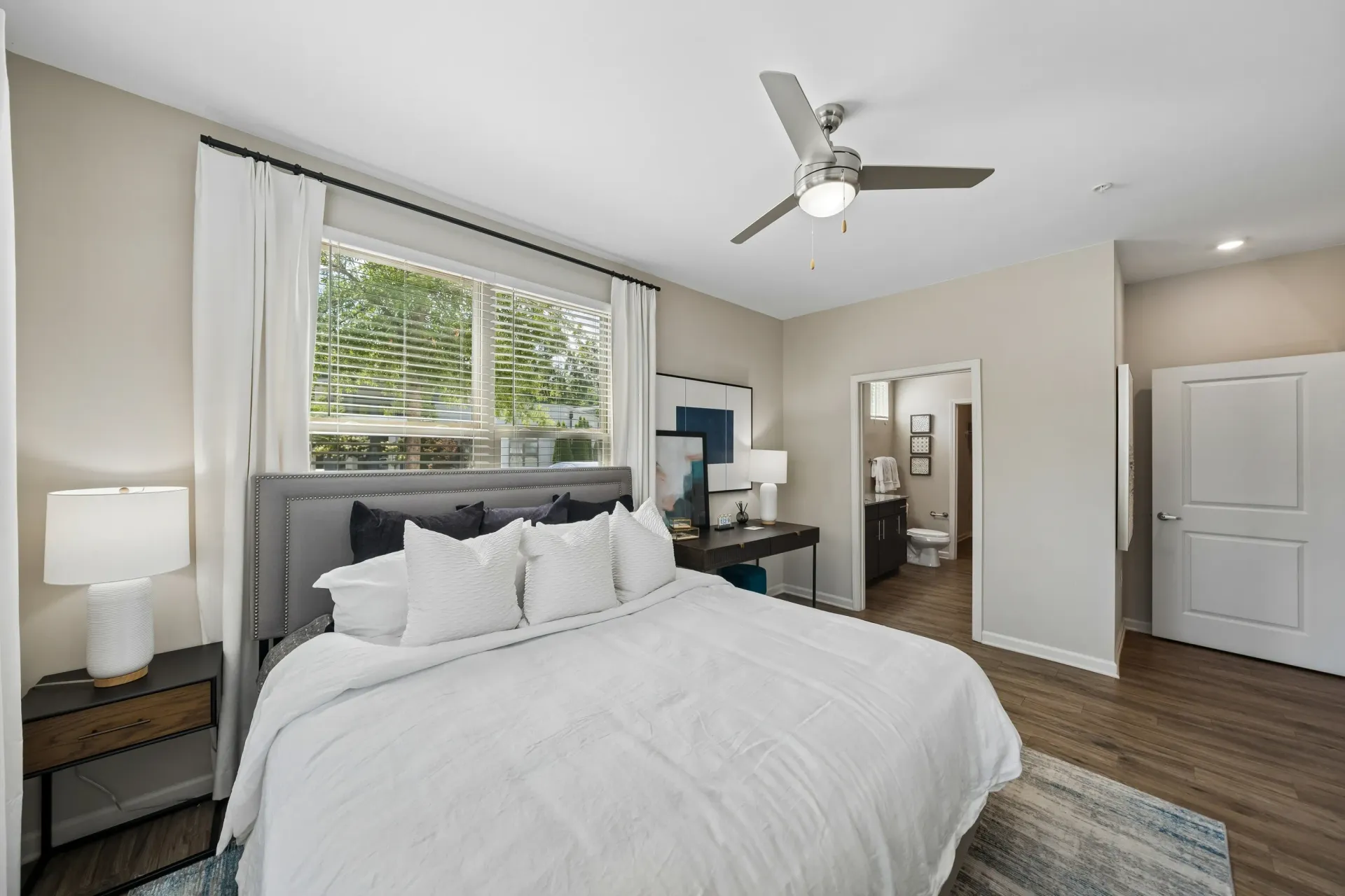 King size bed with white linens and dark accent pillows, nightstand with lamp, desk, and view into ensuite bathroom at Willows at the University in Charlotte, NC.