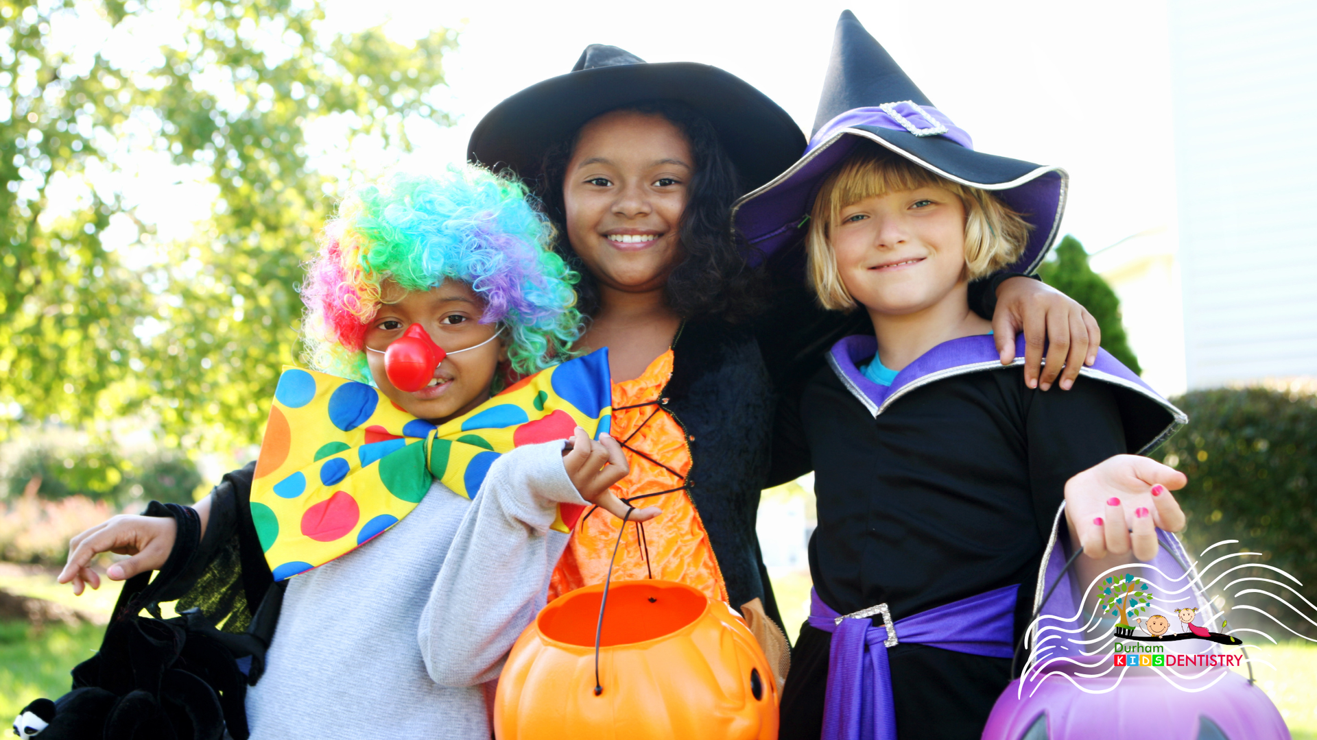 Three children in Halloween costumes: clown, witch, and witch, smiling with candy buckets.