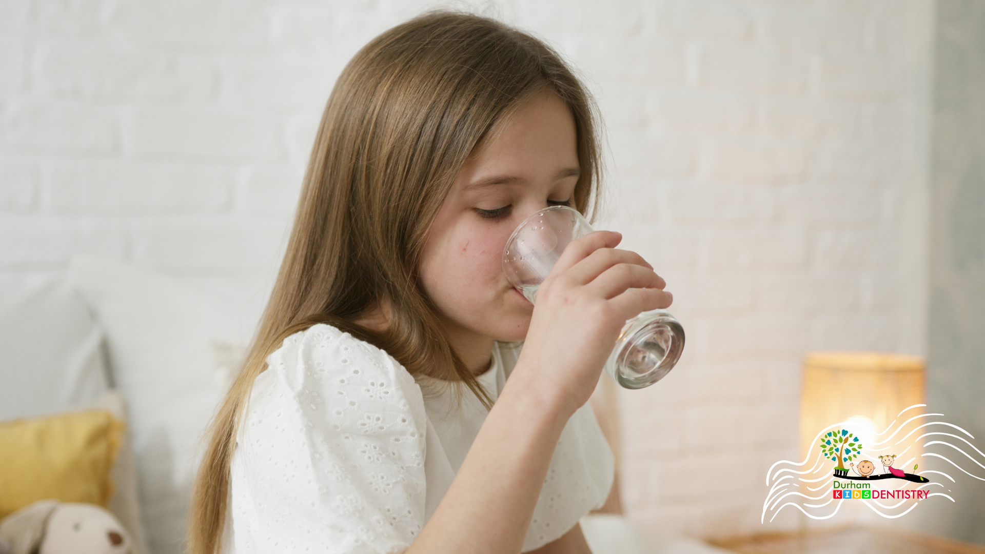 A little girl is drinking water from a glass while sitting on a bed.