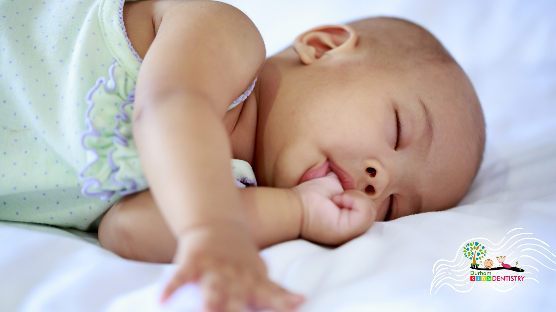 A baby is sleeping on a bed with his hand in his mouth.