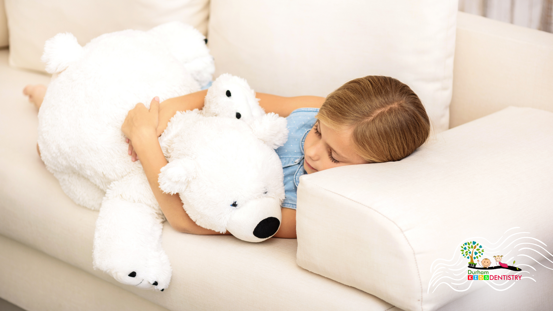 A little girl is laying on a couch holding a large white teddy bear.