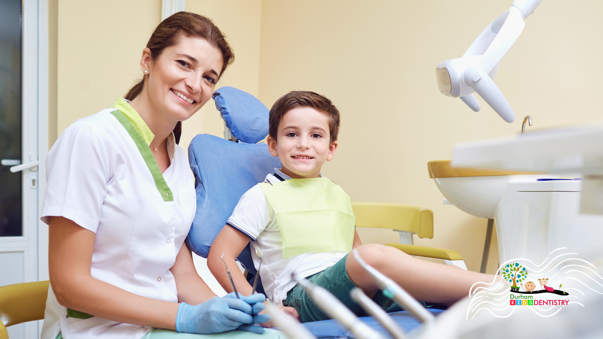 A female dentist is sitting next to a young boy in a dental chair.