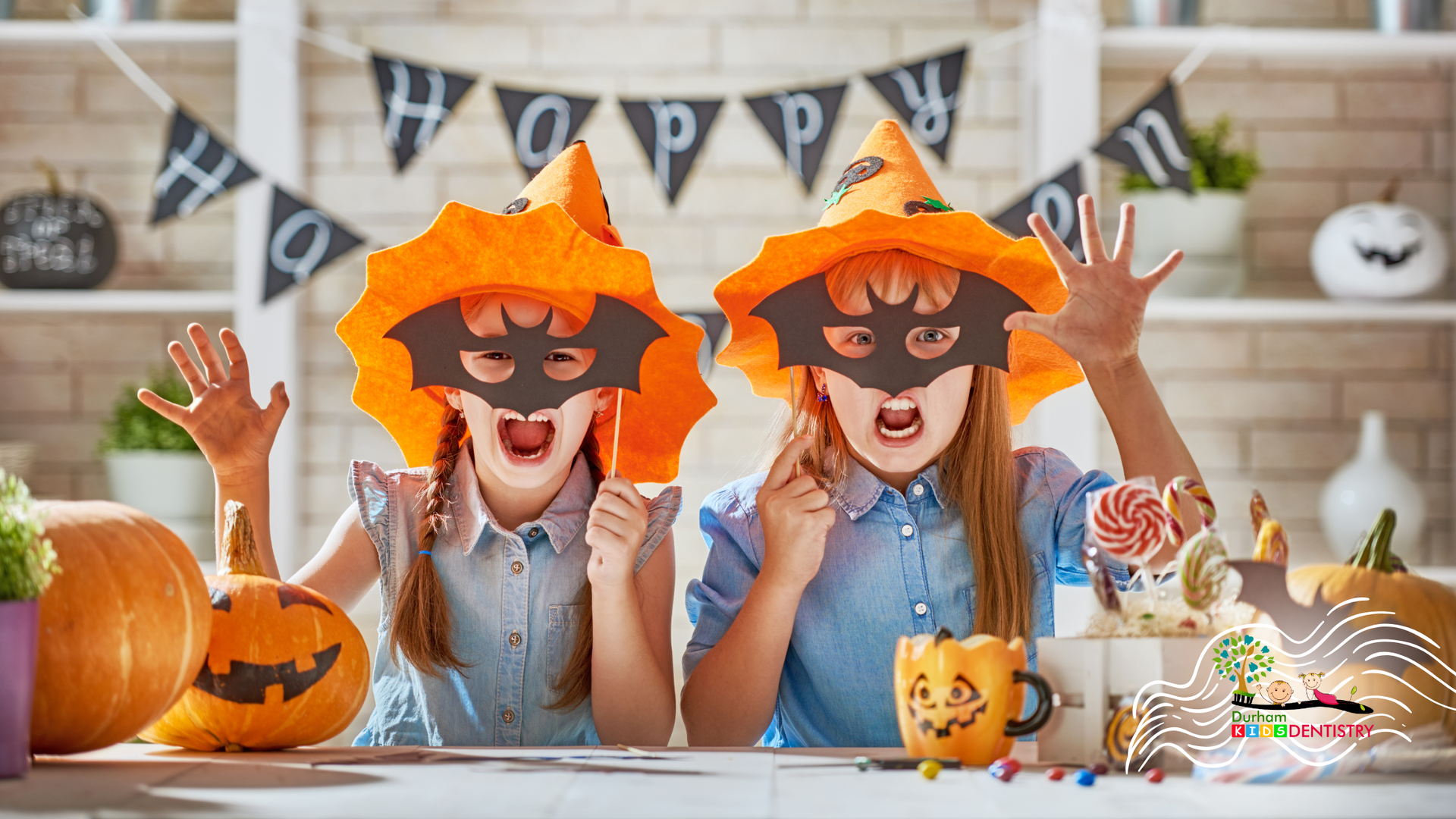Two children in orange pumpkin hats and masks sit at a Halloween table with candy and decorations.