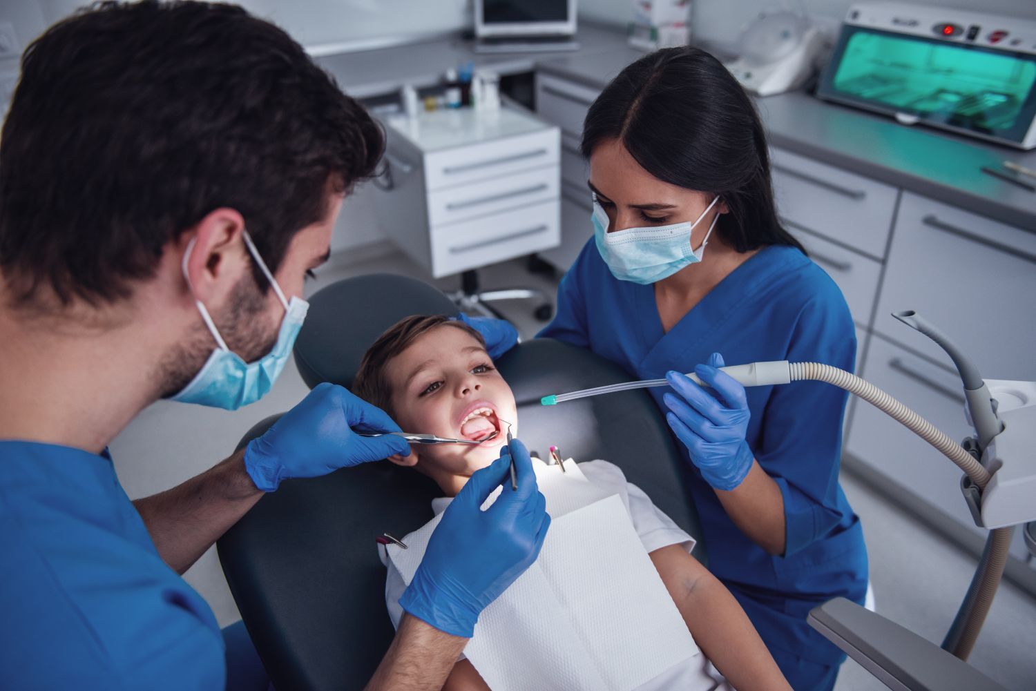 Dentist and assistant examining a smiling child in a dental chair, both wearing masks and gloves