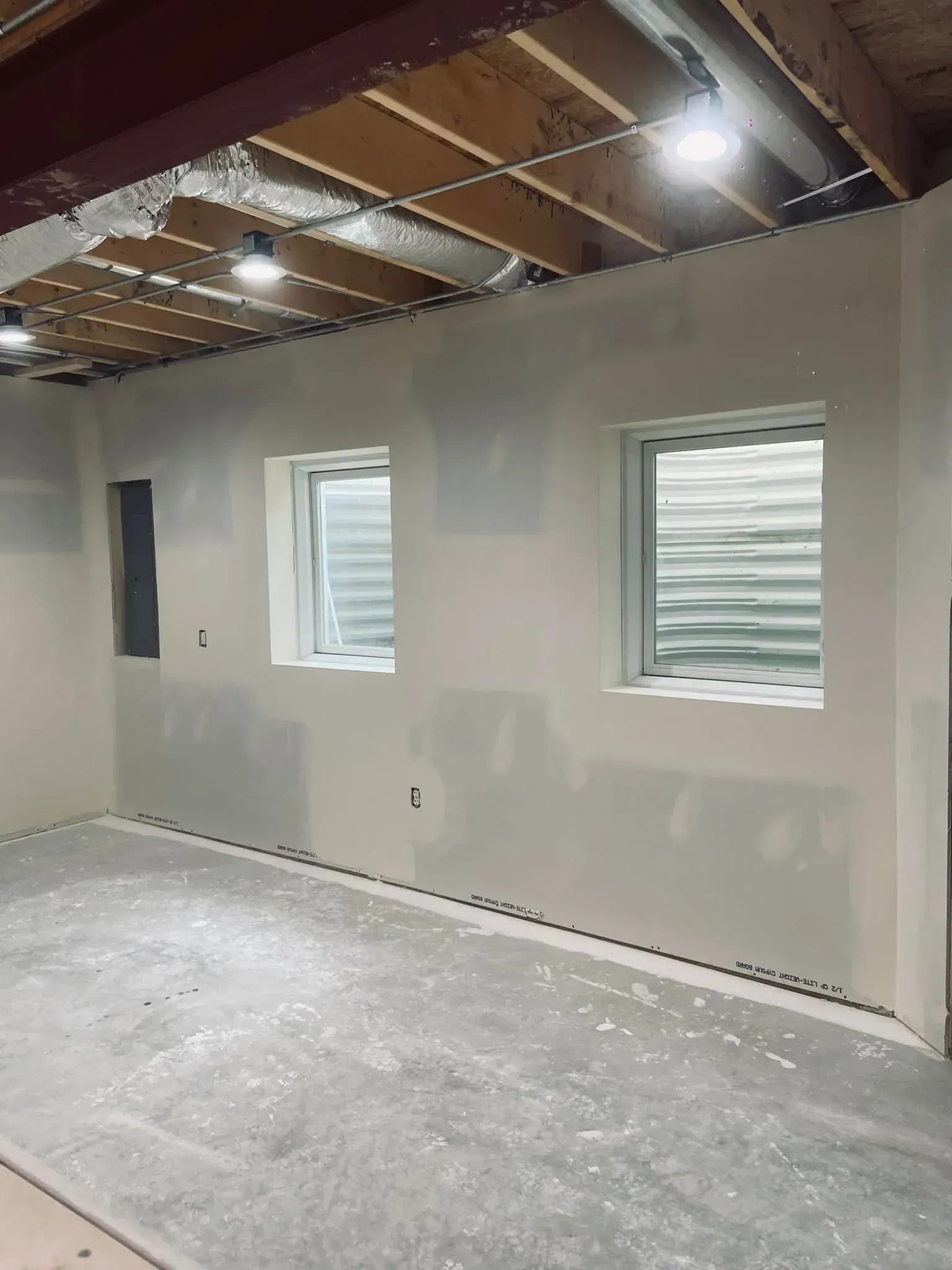 Unfinished basement room with drywall, two small windows, exposed ceiling joists, and a bare concrete floor
