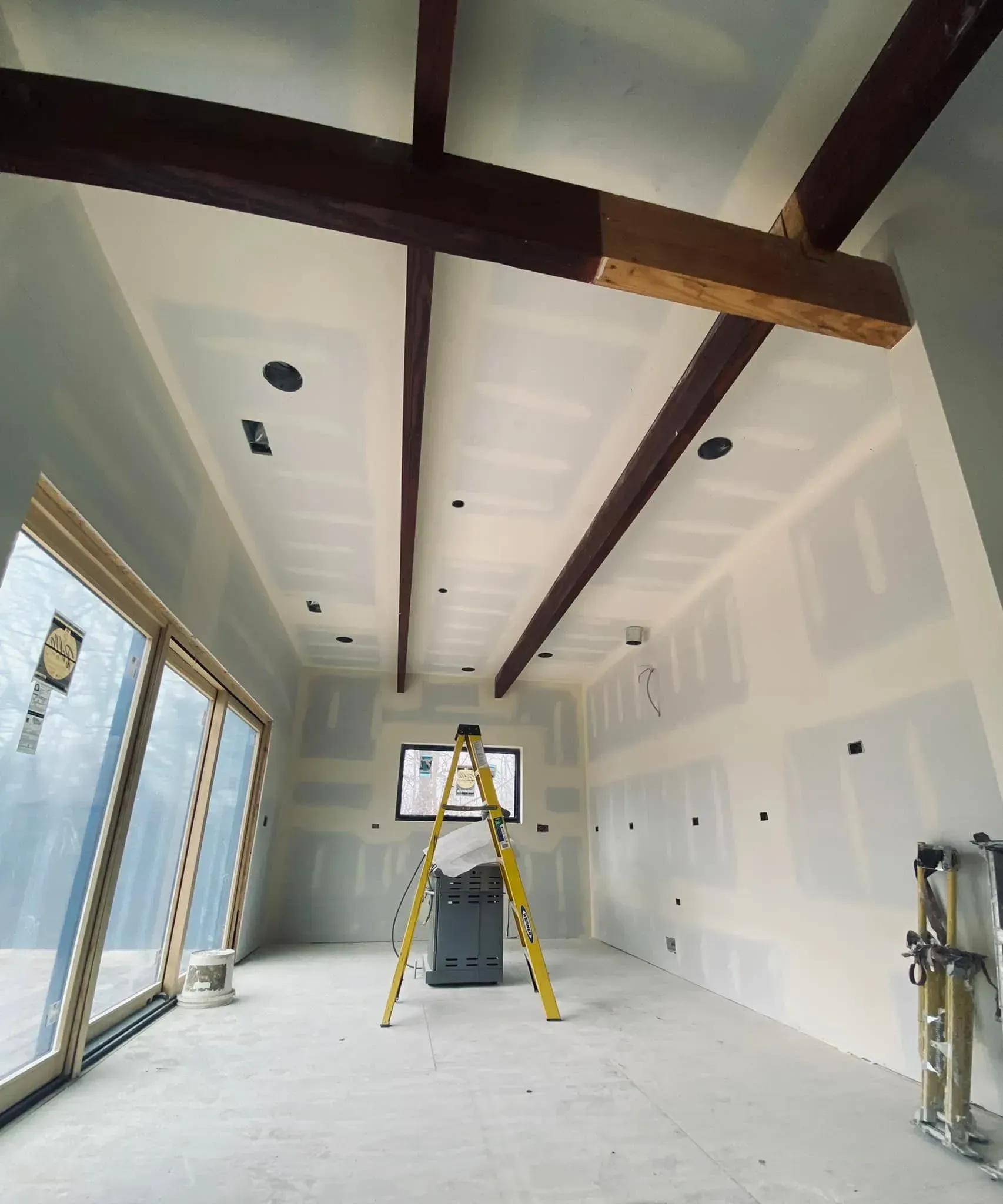Bright unfinished room under construction with exposed beams, ladder, and large windows.