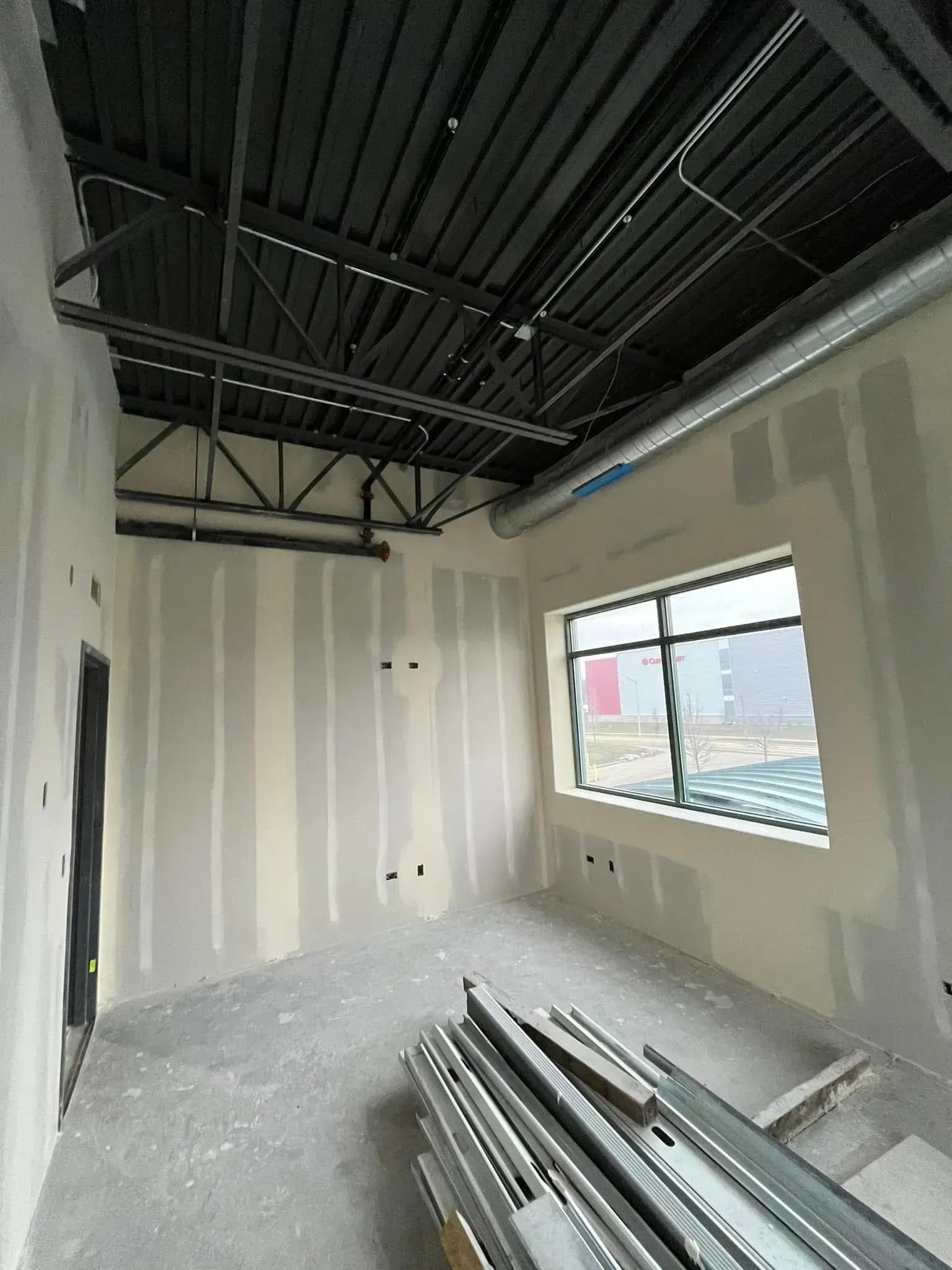 Partially finished room with exposed black ceiling, white walls, window, and stacked metal pipes on the floor
