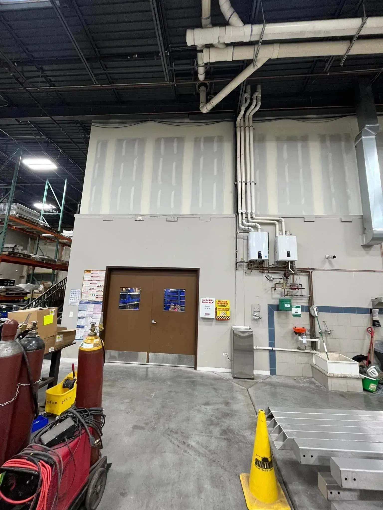 Warehouse interior with tall gray wall, brown double doors, pipes overhead, and industrial equipment.