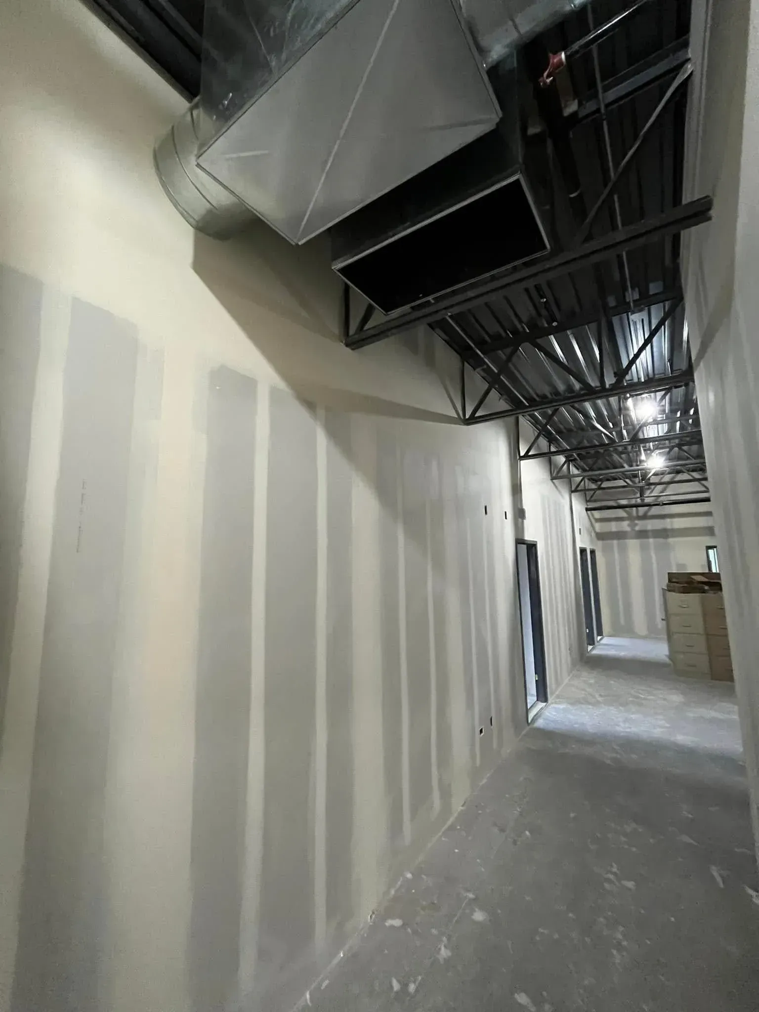 Narrow unfinished corridor with drywall, exposed ducts, and hanging ceiling fixtures under construction