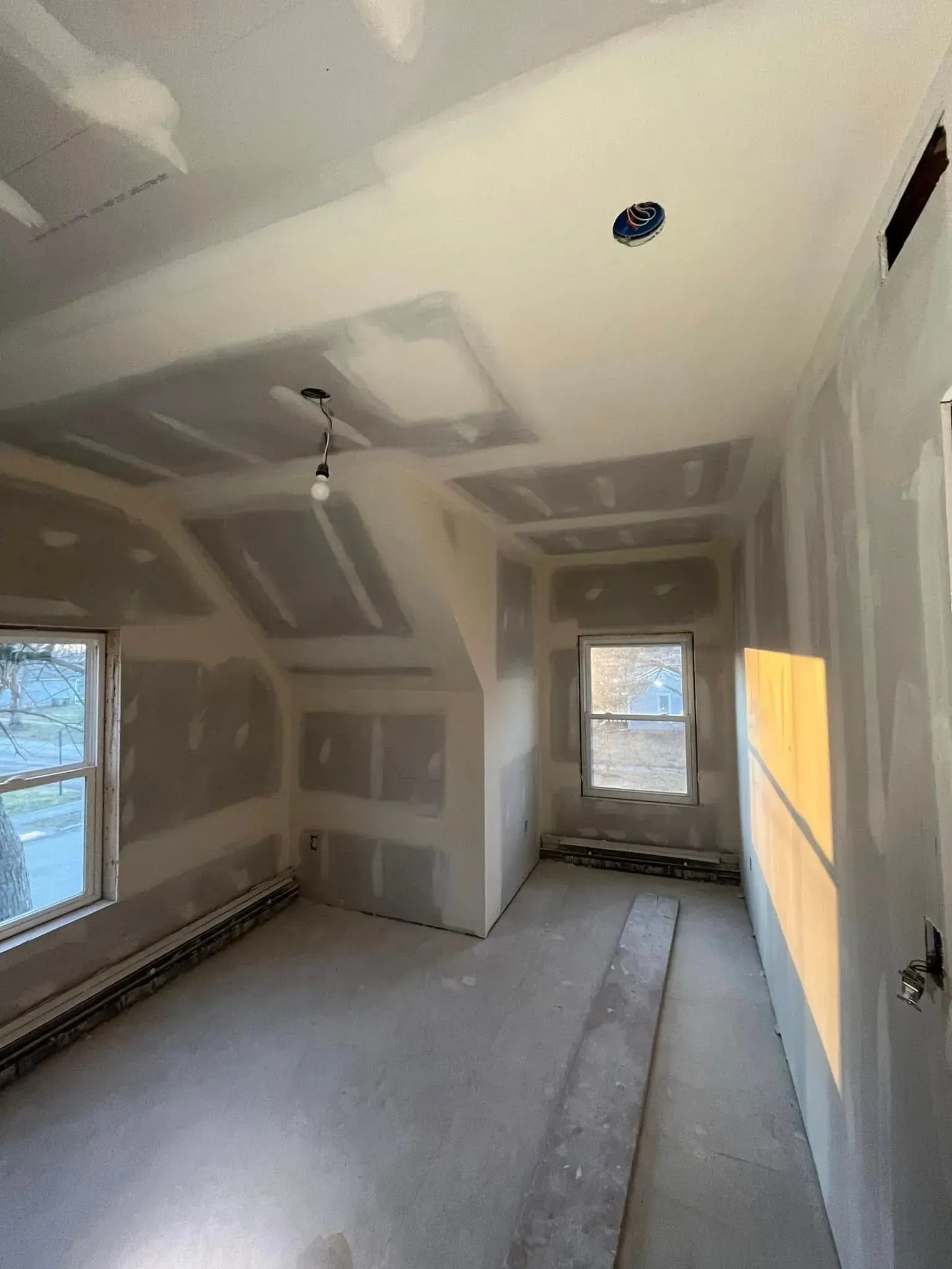 Unfinished drywall room with sloped ceiling, windows, and a doorway to another room